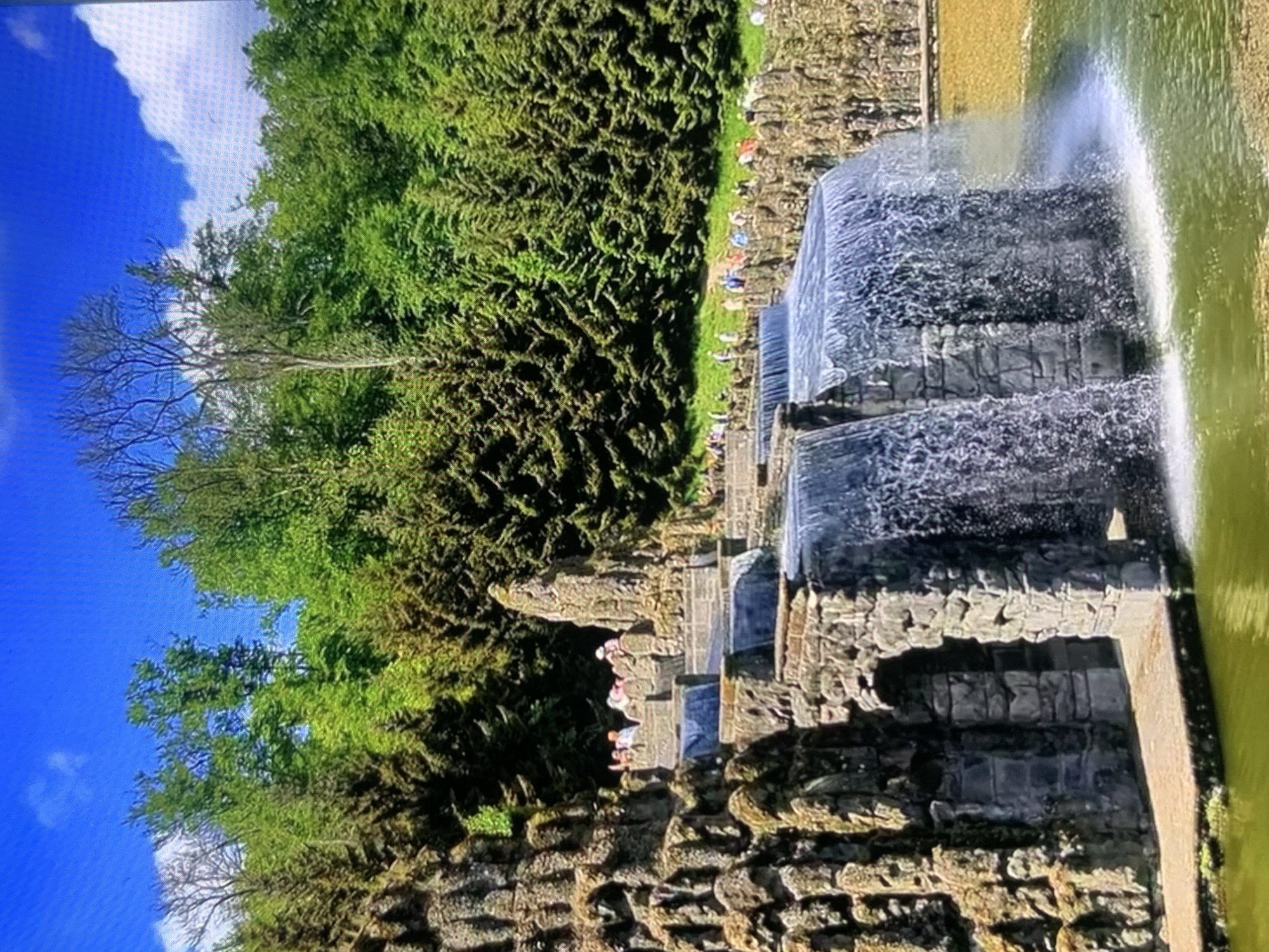 The image depicts a large, multi-tiered artificial waterfall constructed with rough-hewn stones and rock formations. Water cascades down several levels, creating a dynamic and lush environment. In the background, a dense forest of evergreen trees rises under a bright blue sky with scattered white clouds. People are visible on various ledges and pathways surrounding the waterfall, observing the scene. The overall impression is that of a grand garden or park with a significant water feature. The location is not Stavropol, Russia.