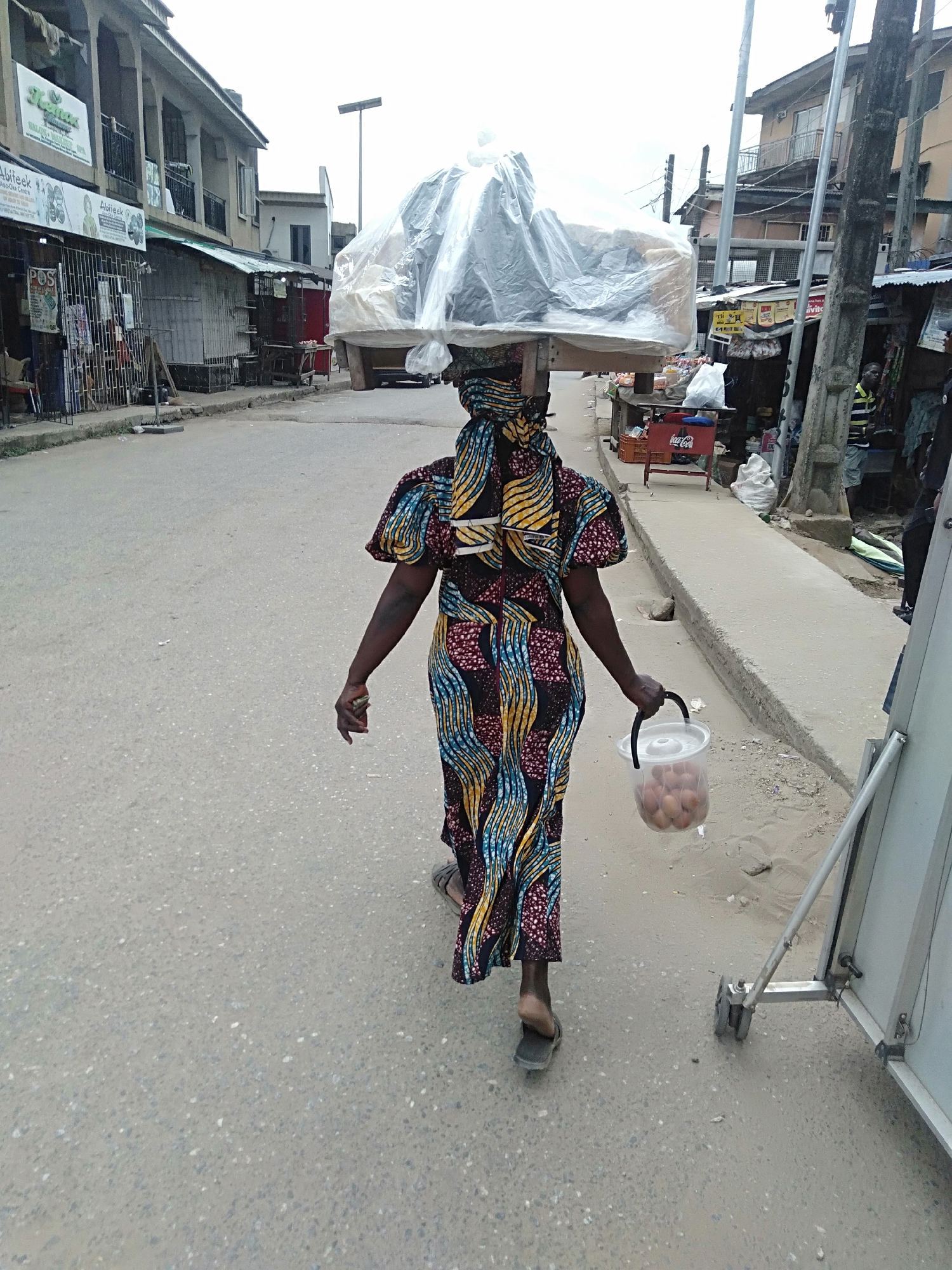 A woman walks down a street in Shangisha, Nigeria, carrying a large, wrapped load on her head and a bucket of eggs in her hand. She wears a long, colorful dress and simple sandals. The street is paved and lined with buildings and shops, some with open fronts. A Coca-Cola cart and a sandwich board are visible near the sidewalk. The sky is overcast.