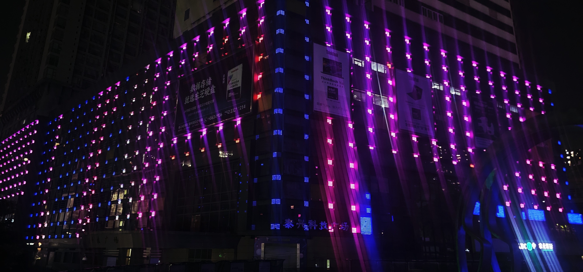 This is a night-time, low-angle, wide shot of a modern building facade in Jiulongpo District, China, adorned with a vibrant LED light display. The building's architecture is obscured by the overwhelming presence of the illuminated lights, which form intricate patterns across its surface. The lights are primarily in shades of purple and blue, with some red accents, and are arranged in a grid-like fashion, with individual lights appearing as small, bright squares. Streaks of light extend outward from many of the LEDs, suggesting a long exposure photograph or movement of the lights. Several large advertisements or posters are visible on the building's facade, partially covered by the LED display. One poster prominently features the "ThinkBook" logo and text in Chinese. The lower portion of the building shows windows and architectural details, also partially illuminated by the external lights. The scene is characteristic of urban nighttime illumination and advertising in a commercial or business district.