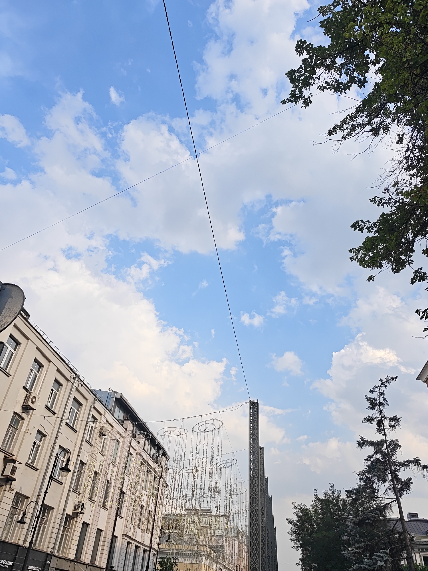The image depicts a view looking up at a city street, likely in Moscow, Russia, with buildings and a clear blue sky dotted with white clouds. The perspective is tilted, suggesting the photo was taken from ground level looking upwards.
In the foreground, on the left, is a tall, light-colored building with multiple windows and air conditioning units. The building's facade is detailed with architectural elements and a black railing along the top. A dark lamppost stands in front of the building.
Further down the street, decorative string lights hang across the width of the road, creating a festive or illuminated atmosphere. These lights are suspended from tall, ornate metal structures that resemble towers or pylons.
On the right side of the image, a tall, slender evergreen tree with dark foliage reaches towards the sky. Behind the tree, more buildings and rooftops are visible, with some having dark, sloped roofs.
The sky is a dominant feature, filled with scattered white cumulus clouds against a vibrant blue background. Two thin, dark wires or cables cross diagonally through the sky, intersecting in the upper portion of the frame. These could be utility lines or part of a decorative lighting system.
The overall impression is of an urban environment, potentially a commercial or pedestrian street, decorated for an event or season, under a bright, partly cloudy sky. The tilt of the image adds a dynamic and upward-looking quality to the scene.