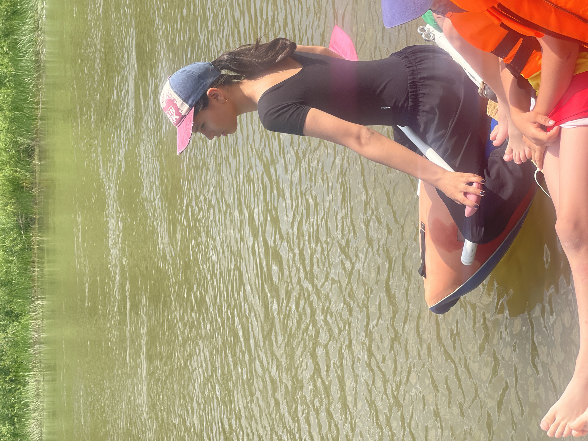 A young person wearing a black one-piece bathing suit and a baseball cap sits on the edge of a colorful inflatable raft in a river. The raft is orange with blue trim. The person is looking to the right, with a paddle resting on their lap. The river is murky green and reflects the bright sky. In the background, a lush green embankment lined with tall grasses borders the river. Another person, wearing an orange life vest and a purple hat, is also visible on the raft. The scene appears to be a warm, sunny day. The city of Nezhinka, Russia is not depicted in this image.