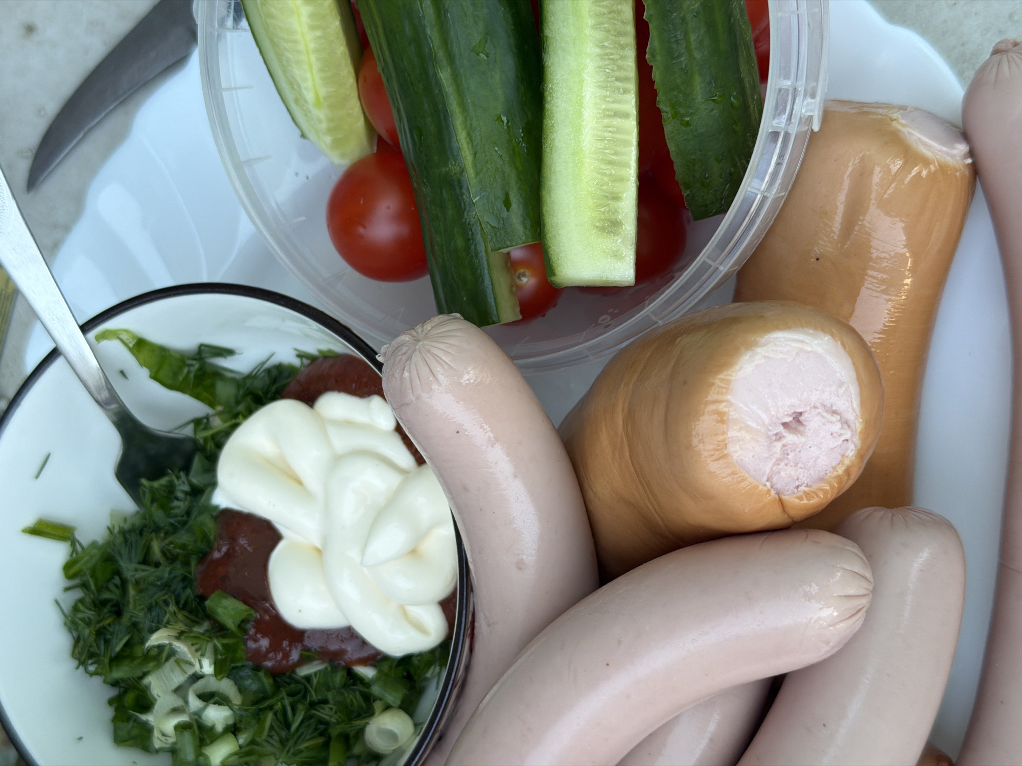 The image shows a plate with food, likely a picnic or outdoor meal. There are several sausages, both pale pink and brown, dominating the right side of the frame. A clear plastic container holds cucumber spears and small red tomatoes. A white bowl contains chopped greens (likely dill and onions) with blobs of red sauce and mayonnaise. A fork rests in the bowl. A knife is partially visible at the upper left edge of the image. The location is described as Chaikovsky, Russia.