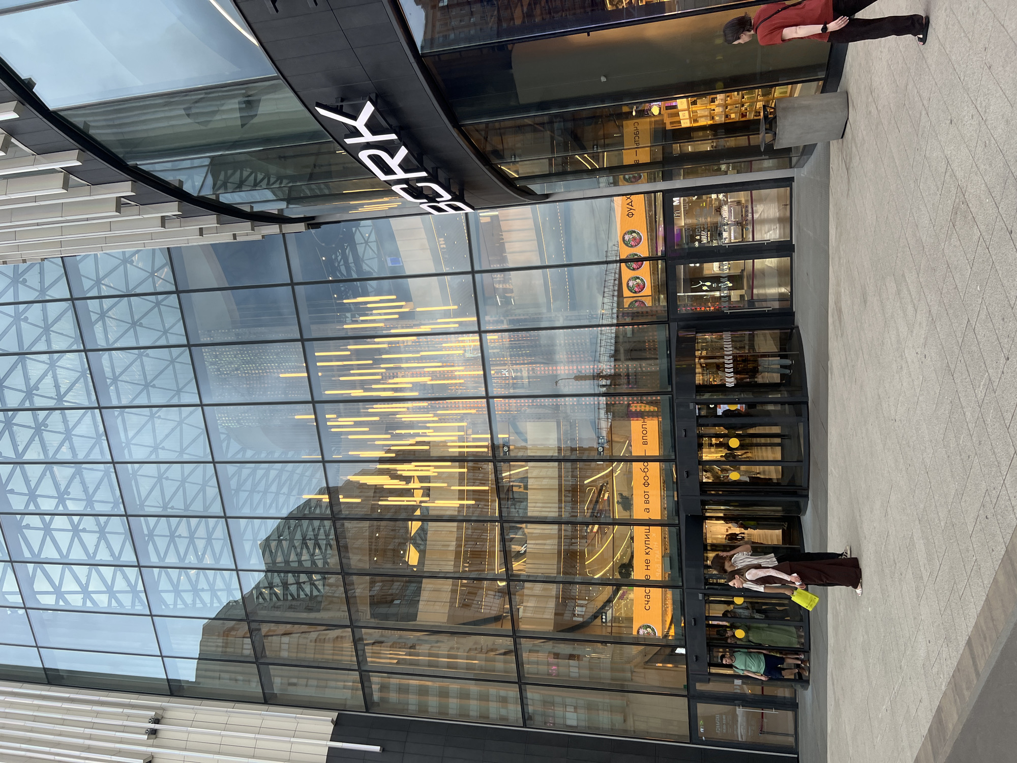 This image depicts the exterior of the Galeria Shopping Mall in Saint Petersburg, Russia. The prominent feature is a vast, multi-story modern building with an extensive glass facade composed of numerous rectangular panels framed in black. Through the glass, multiple interior levels, escalators, and numerous long, vertical yellow-orange light fixtures hanging from the ceiling are visible. A large, illuminated horizontal sign above the main entrance reads in Russian, "счастье не купишь, а вот фо-бо — вполне" (happiness you can't buy, but pho-bo - quite), with additional text "ФУД" (FOOD) to the right.
To the right of the main glass facade, a curved section of the building, darker in color, features vertical light-toned panels and a vertically aligned, illuminated white "BORK" logo. The ground level in front of the building is a paved pedestrian area made of light grey tiles, with reflections of the sky and surrounding structures visible in the upper portions of the glass facade.
The main entrance consists of multiple revolving glass doors. In the immediate foreground-left, a young woman with dark hair, wearing a white long-sleeved top, dark brown overalls, and white sandals, holds a bright yellow shopping bag. She stands beside a young man wearing a white and grey striped t-shirt and dark trousers. In the foreground-right, a woman with short dark hair, dressed in a dark red t-shirt and dark trousers, walks away from the viewer, looking towards the BORK store. A dark grey concrete trash receptacle is situated on the pavement near her. Several other individuals are visible near the revolving doors, either entering or exiting the building. The sky appears overcast.