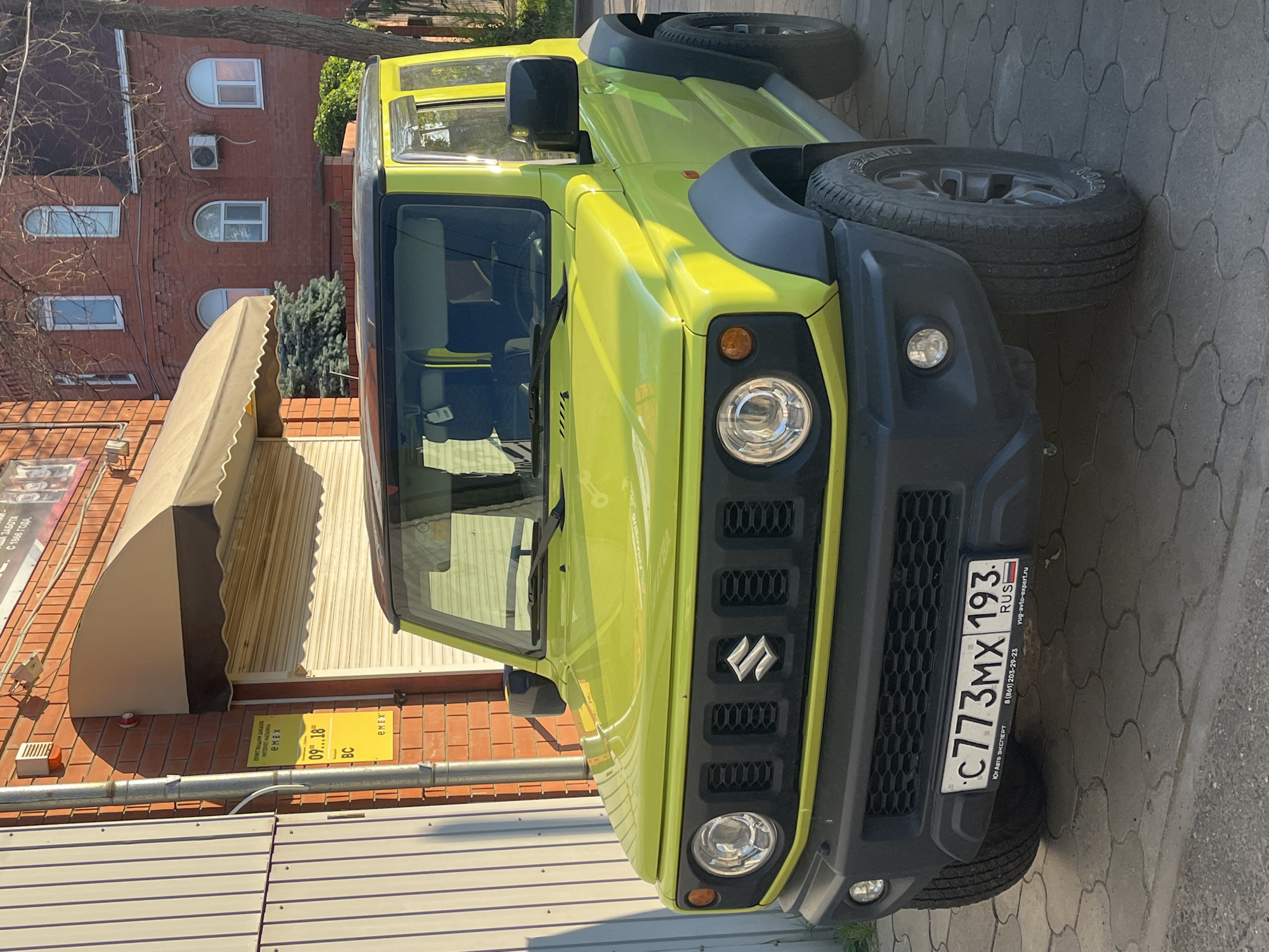 This media file shows a bright yellow Suzuki Jimny parked on a paved surface next to a brick building. The car is the primary subject, with its front grill, headlights, and spare tire prominently displayed. The building appears to be a commercial establishment, with signage indicating a pick-up point for an online store called "EMEX". Text on the sign also provides operating hours (09:00-18:00) and a phone number. Another piece of text on a smaller sign near the car reads "Юг-Авто ЭКСПЕРТ" and a website address "yug-avto-expert.ru". The scene suggests a daytime setting, possibly in the afternoon, given the shadows. The sky is visible in the background, and the weather appears to be clear. The overall atmosphere is neutral, focusing on the vehicle and its surroundings. The image is oriented sideways.