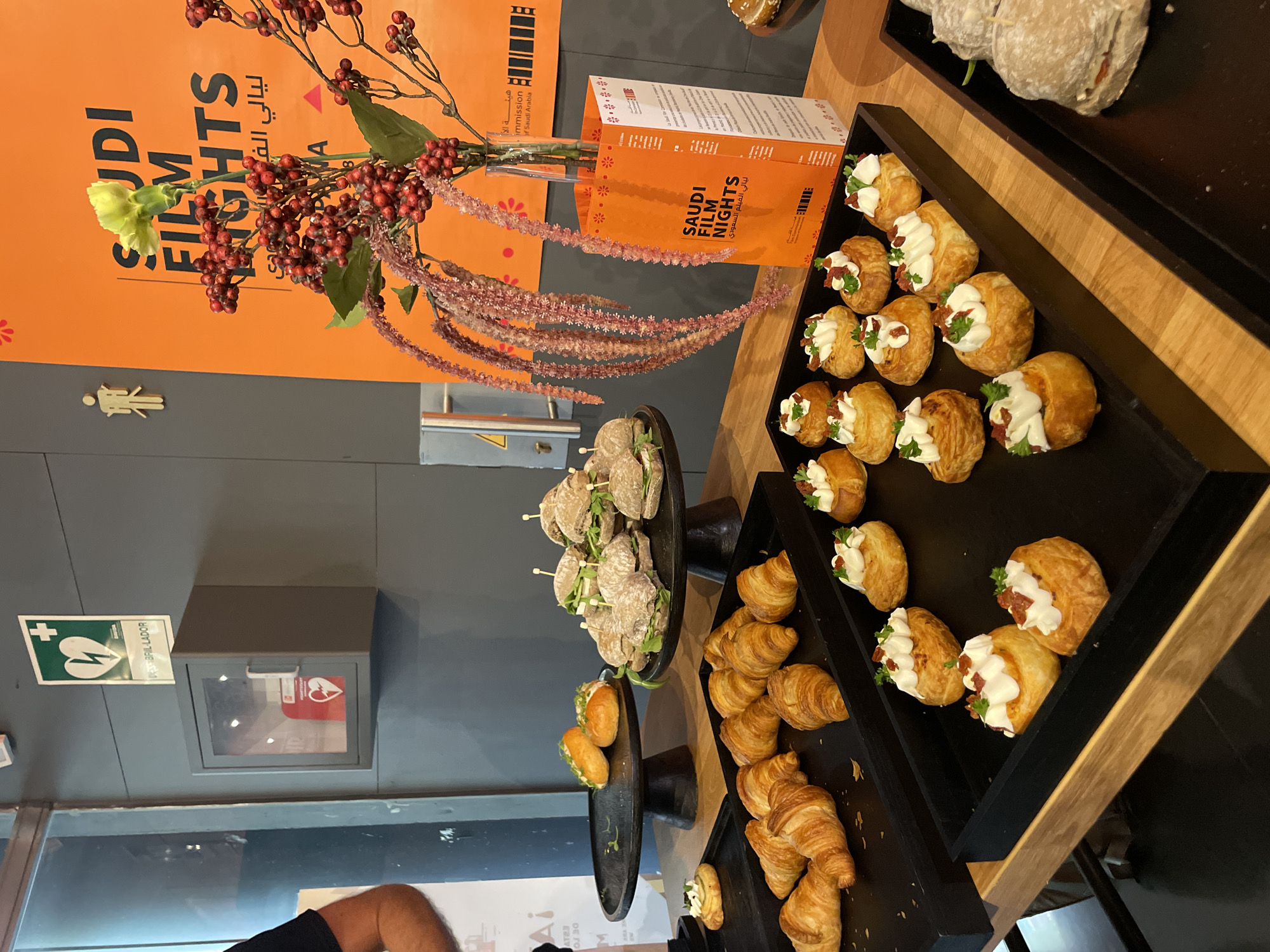 The image depicts a buffet table, possibly in Barcelona, Spain, given the presence of a sign in Catalan related to a defibrillator (the Catalan word for defibrillator is "desfibril·lador"). The table features various pastries and small sandwiches displayed on black trays and stands. In the background, there's an orange poster with "SAUDI FILM NIGHTS" in English and Arabic. A floral arrangement in a vase sits near the poster. The pastries include croissants and puff pastries with cream and toppings. There are also small sandwiches on skewers. A defibrillator cabinet is visible on the wall above the table. A person’s arm and shoulder are visible at the bottom-left corner of the frame.