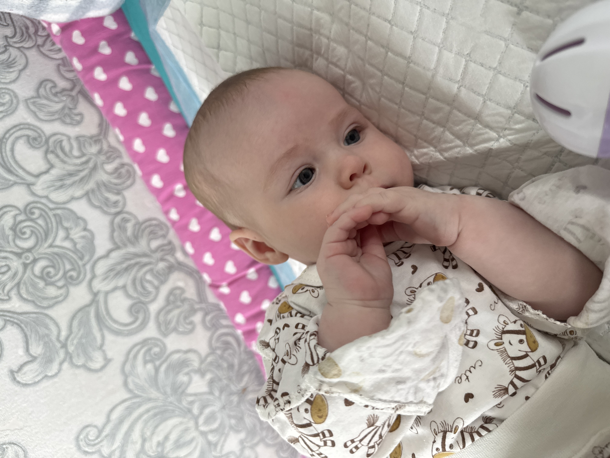 A baby lies on a white quilted surface with a pattern of gray floral designs to the left. The baby has light skin, blue eyes, and is wearing a white patterned onesie with brown zebras and hearts. The baby's hands are near its face. In the background, there is a pink pillow with white hearts and a blue object. A white object with purple stripes is visible in the upper right corner. The location is Кемуль, Russia.