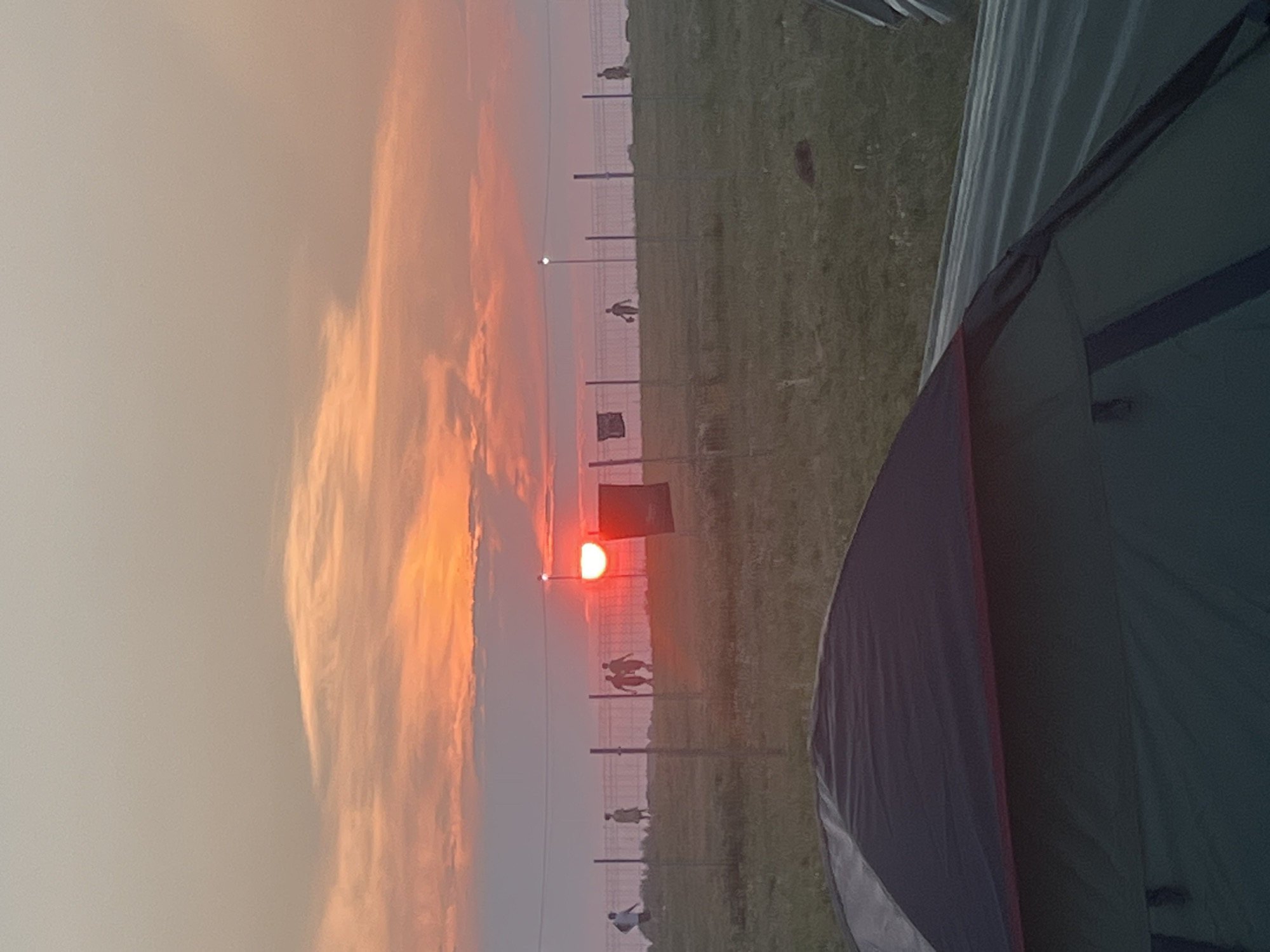 The image depicts a sunset scene with a prominent orange and pink sky. In the foreground, the top of a green and black tent is visible. A metal fence runs horizontally across the middle ground. Beyond the fence, several silhouetted figures are scattered across a grassy field. One figure is walking towards the left, and a pair of figures are walking together towards the center. A bright, round object, likely the sun, is visible near the center-right, appearing as a red disc behind a light pole. Several other light poles are spaced along the fence. A dark rectangular object hangs from the fence on the right side of the sun. The overall environment appears to be an open field, possibly a campsite, with a fence marking a boundary. The location is specified as Surakovo, Russia.