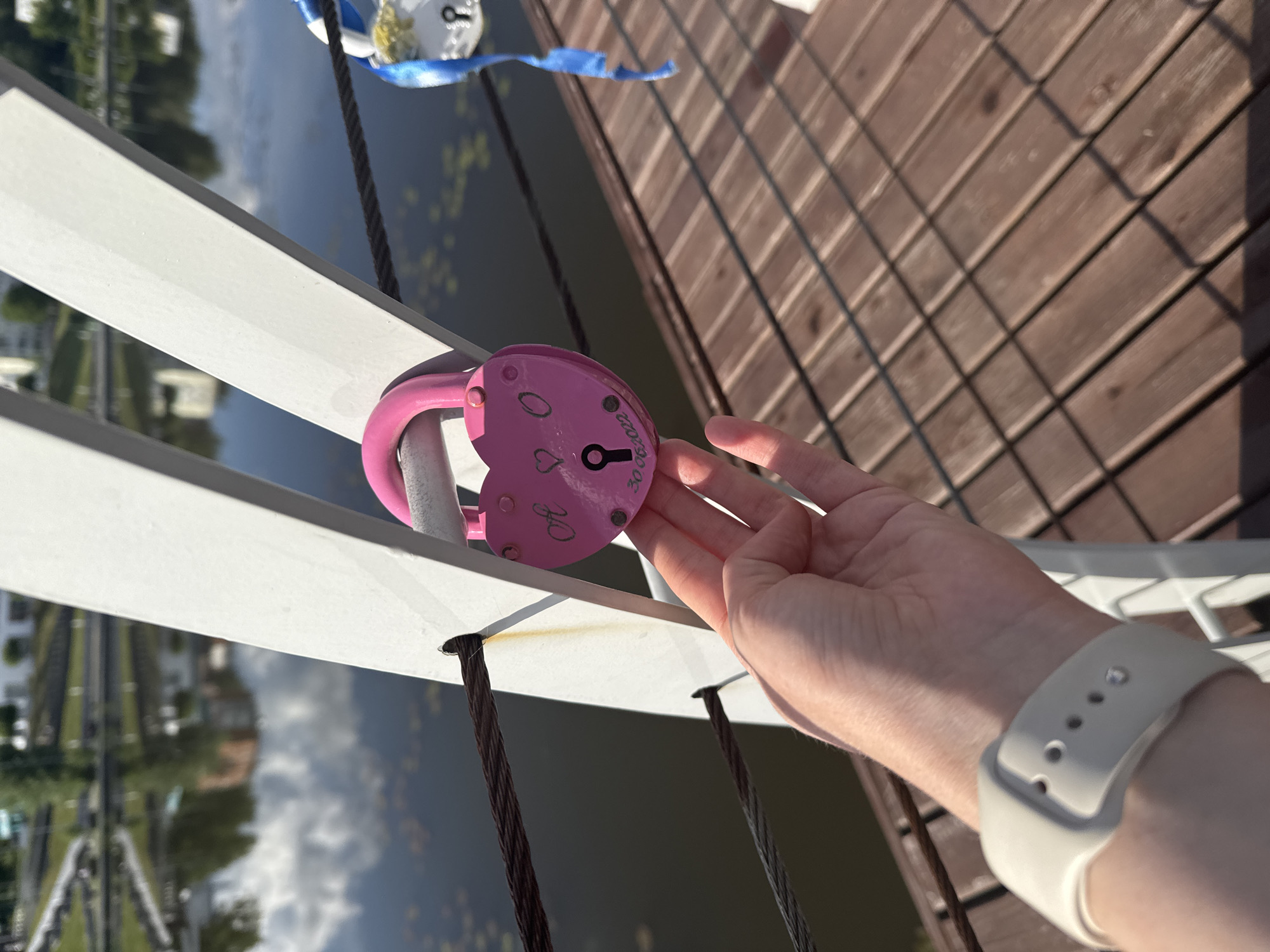 A pink heart-shaped padlock is affixed to a white railing on a bridge. A hand reaches towards the lock. The railing overlooks a body of water and some foliage. The bridge has a wooden deck. The city of Votkinsk, Russia, is visible in the background. The person is wearing a white wristband.