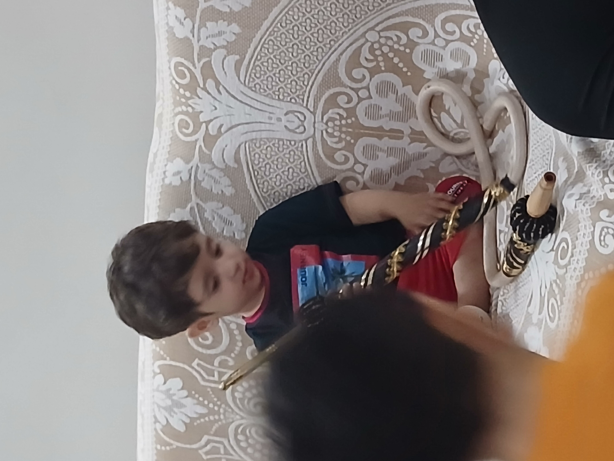 A young boy with brown hair is sitting on a beige and white patterned couch, holding a long, dark hose with gold accents. He is wearing a black t-shirt with a red and blue graphic that reads "JOURNEY" and red shorts. In front of him on the couch are two coiled white hoses and another hose with a black and gold patterned handle. The couch has an intricate, ornate white pattern against a beige background. The wall behind the couch is a plain, light color. In the foreground, the back of a person's head, with dark hair, is visible on the left, and the shoulder of someone wearing a black garment is visible on the right. The scene appears to be indoors. The exact location in Barza Municipality, Syria, cannot be determined from the image.