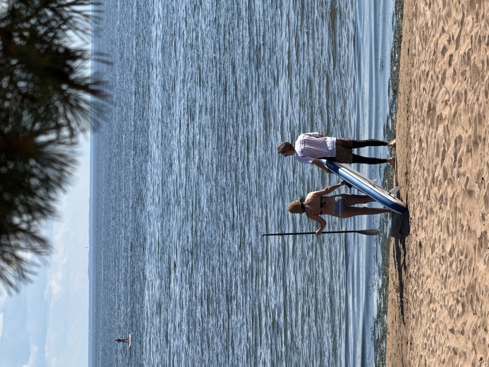 Two adults, a woman and a man, stand on a sandy beach facing the water, ready to paddleboard. The woman, on the left, wears a bikini and a straw hat and holds a paddle. The man, on the right, wears a t-shirt, shorts, and leggings, and holds a paddleboard. The water is blue and rippling, with a few other paddleboarders visible in the distance. The sky is partly cloudy. The scene is located in Zelenaya Roshcha, Russia, likely on a beach along the coast.