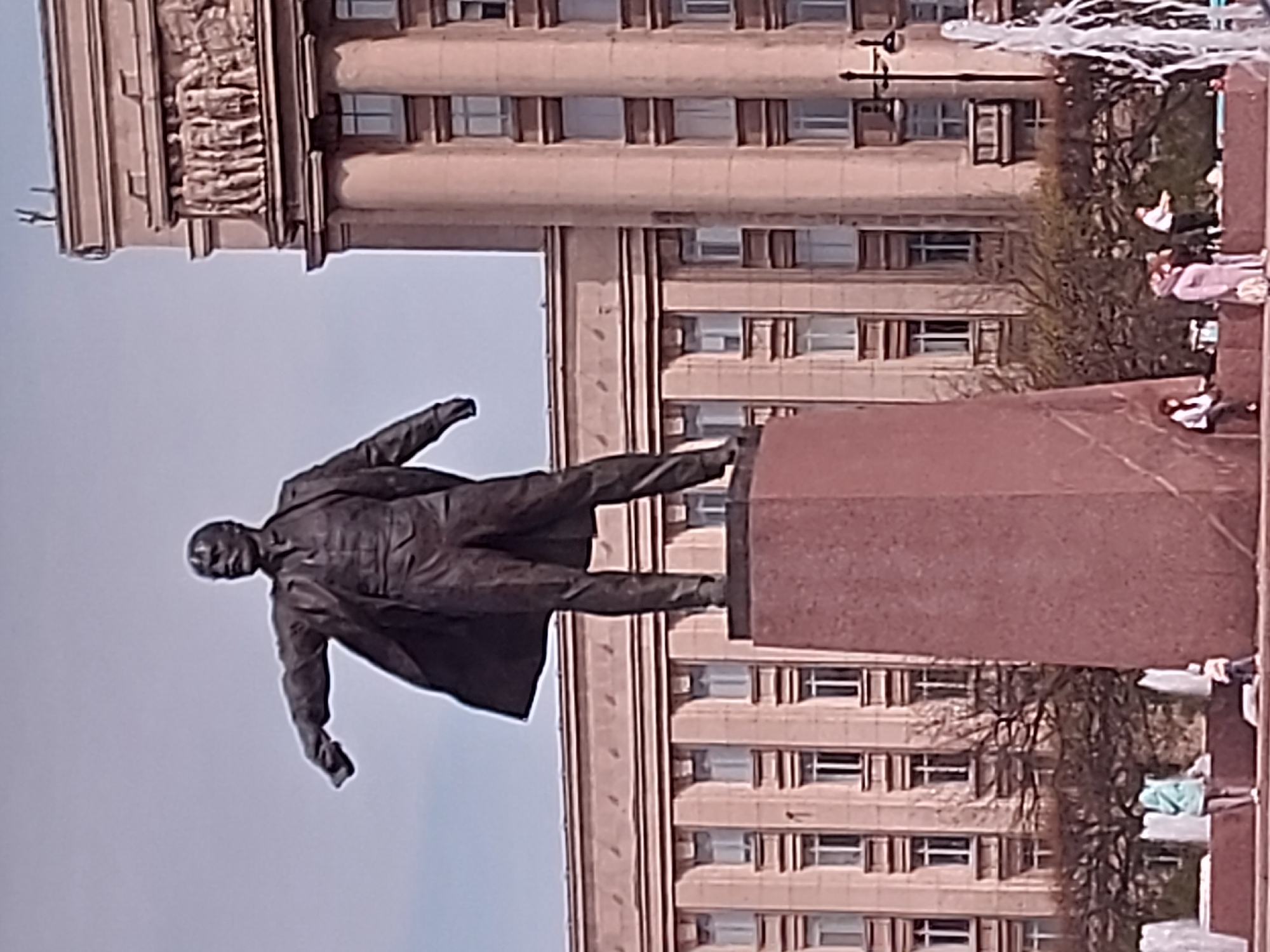 The image shows a statue of Lenin in front of a Stalinist building, likely located in Saint Petersburg, Russia. The statue is in the traditional pose of Lenin, with one arm outstretched and the other at his side, atop a tall plinth. The building in the background is a large, multi-story structure with a symmetrical facade and many windows, characteristic of Stalinist architecture. People are visible in the background.