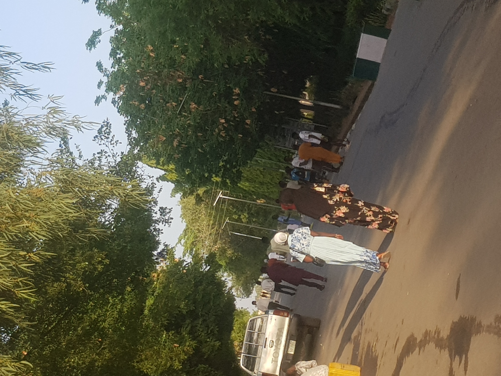 The image shows a street scene in Dabawa, Nigeria. A group of people are walking along a paved road, with trees lining both sides. A white Toyota pickup truck is parked on the side of the road, and at least ten people are walking along the street. Some of the people are wearing long dresses, and others are wearing more casual clothing. The road is gray, and the sky is visible between the trees.
