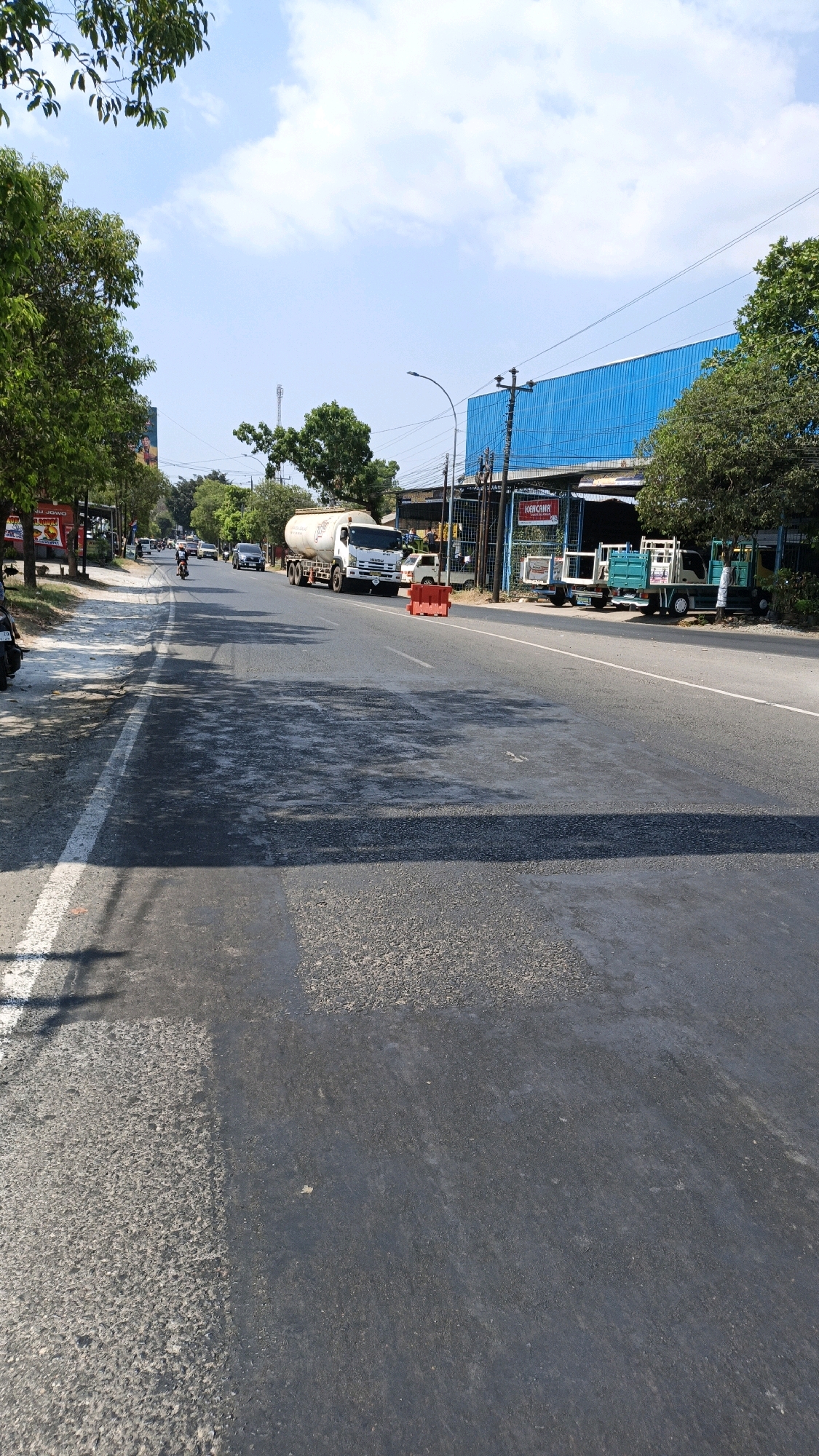 The image depicts a street scene in Sumbring, Indonesia. The road surface shows signs of repair, with patches of new asphalt laid over older sections, creating a textured appearance. A white line marks the edge of the lane.
On the right side of the road, a bright blue building with a corrugated metal facade is visible. Several vehicles are parked or driving in front of it, including a white tanker truck, a white car, and two light-colored trucks with teal-colored beds. A red construction barrier is placed in the middle of the road.
To the left of the road, trees with green foliage line the sidewalk. The sky is blue with scattered white clouds. In the distance, a cell tower is visible. The overall scene suggests an active, possibly commercial area.