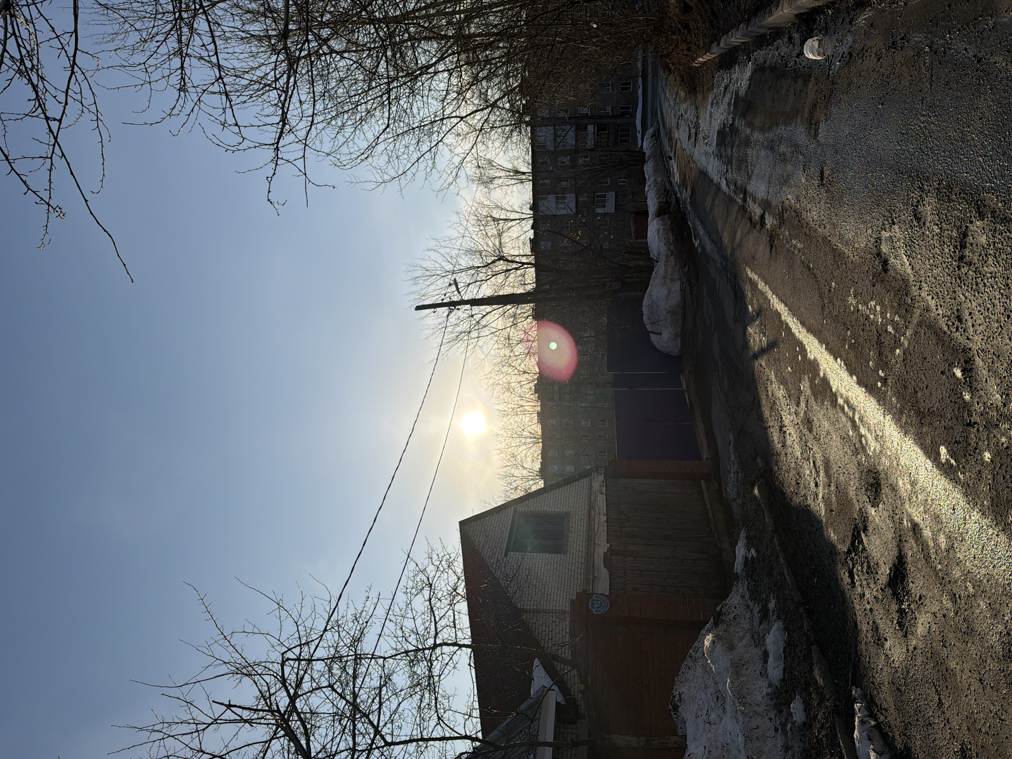 The image shows a street in Berezniki, Russia, on a sunny day. A narrow asphalt road runs along the right side of the frame, showing patches of melting snow and ice. A residential building is visible in the background. The sky is clear, with a bright sun. Bare tree branches frame the top and left edges of the photo. A small brick and wood building is along the road. Overhead power lines are also visible.