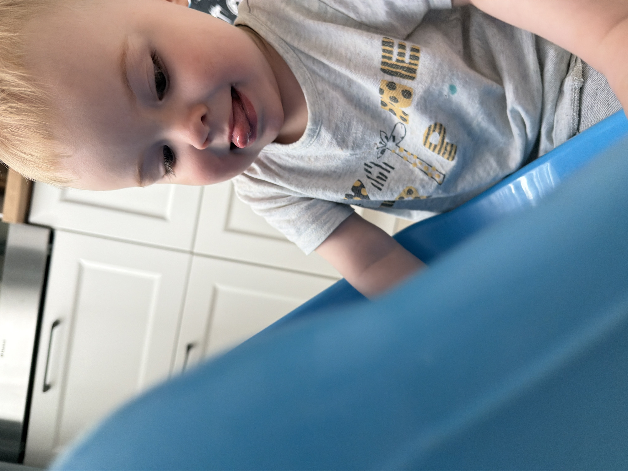 A young child with blonde hair and light skin, wearing a light grey t-shirt with a giraffe and letter patterns on it, is sticking their tongue out. The child is positioned close to a blue plastic object, possibly a container or toy. In the background, there are white kitchen cabinets and a stainless-steel appliance. There is no indication of the location being in Votkinsk, Russia.