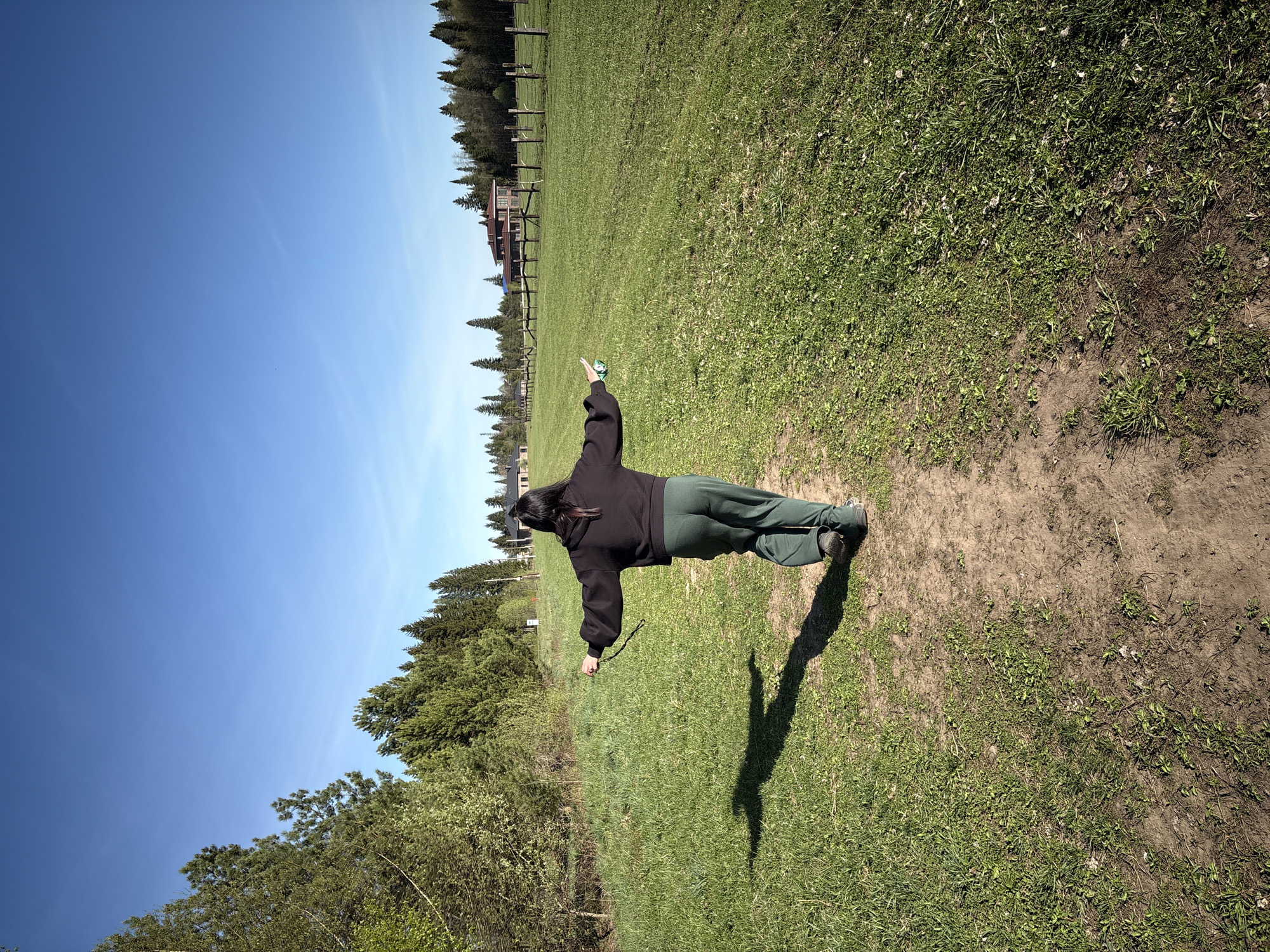 A woman stands in a grassy field in Азинский, Russia. She wears a black hooded sweatshirt and green pants. Her arms are outstretched, forming a T-shape. A dirt path stretches behind her. The sky is bright blue with some trees in the distance. A fence and a building are visible farther away.
