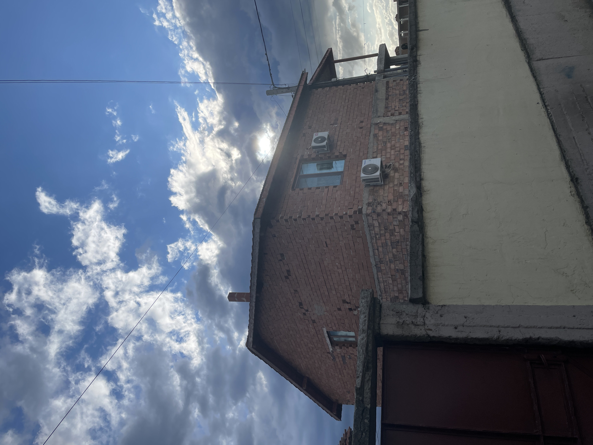 The image displays a partial view of a building in Ihtiman, Bulgaria, under a dramatic, partly cloudy sky. The building is constructed with two distinct sections: one primarily of exposed red brickwork, and another adjacent part finished with a light cream or yellow plaster.
On the brick section of the building, which includes a visible gable roofline, two white outdoor air conditioning units are mounted. A rectangular window is also visible on this wall, reflecting the bright sky. Concrete structural elements, including a beam above a dark red gate or door, are present in the lower right portion of the frame, indicating an entrance or boundary.
The sky dominates the upper and left parts of the image, showcasing a mix of clear blue patches and various cloud formations. Some clouds are bright white and fluffy, while others are darker and more voluminous, suggesting dynamic weather. The sun is prominently visible in the upper center, shining brightly through or from behind clouds, creating a distinct sun flare. Thin black power lines or cables traverse the clear blue sky horizontally in the upper left.
The scene depicts an ordinary outdoor setting with no people or specific activities taking place. The bright sunlight indicates it is daytime, likely mid-day or afternoon. No visible text is present in the image.