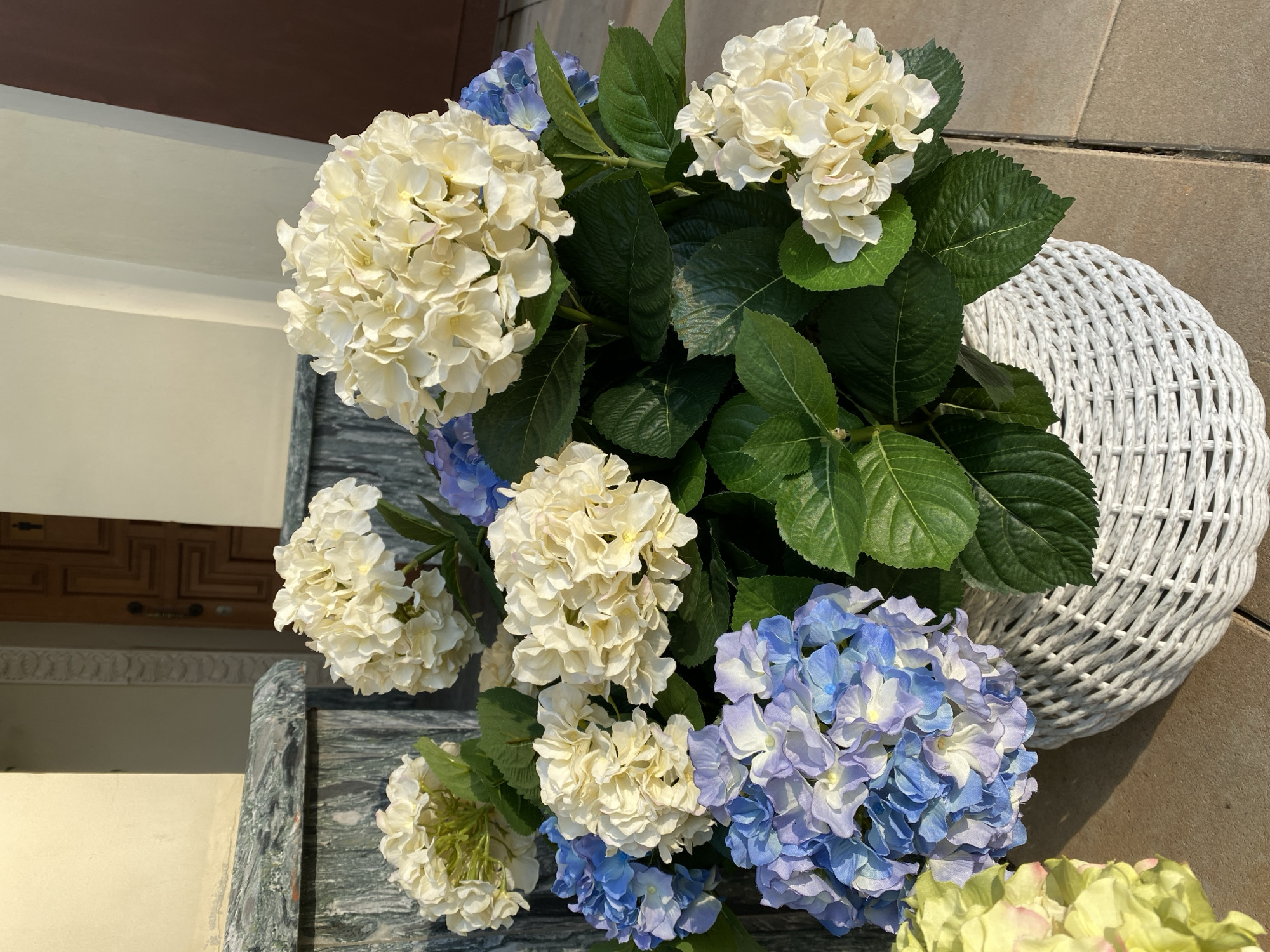 A high-angle, eye-level shot displays a floral arrangement with blue and white hydrangeas and green leaves. The arrangement sits partially on a dark marble surface to the left and a light brown tiled surface to the right. A white woven basket is visible to the right of the flowers. The background shows a building exterior with a wooden door featuring geometric patterns. Based on the architectural style and door design, a reasonable interpretation would place this in a location similar to one found in Yekaterinburg, Russia, known for its diverse architectural styles. However, there are no other distinctive elements that could confirm the exact location of the image.