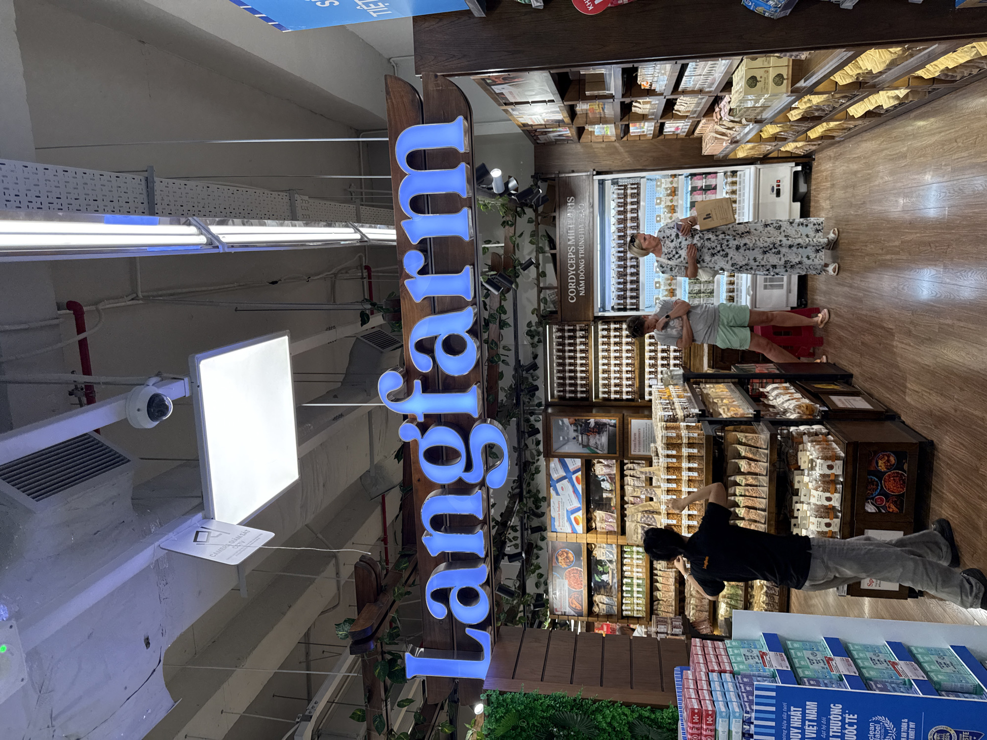 The image shows the interior of a store named "Langfarm" in Nha Trang, Vietnam. A large blue neon sign that spells "Langfarm" is prominently displayed above a wooden structure decorated with artificial greenery. In the foreground, a person wearing a black shirt and grey pants is seen from behind, facing shelves stocked with various products. To the right of this person, two women are standing and looking at products on display. One woman, wearing a white and black floral dress, holds a shopping bag. The other woman, in a grey t-shirt and green shorts, has her hand on her chin. The store appears to sell food items, with shelves filled with packaged goods and jars. The lighting is bright, and the store floor is made of wood-patterned laminate. A security camera is visible on the ceiling.