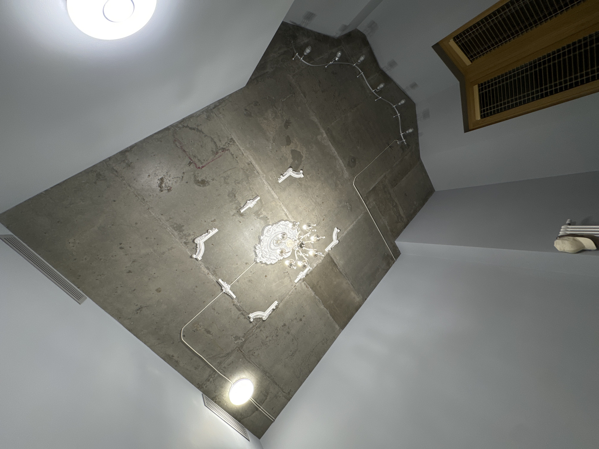 A low-angle interior shot shows a ceiling with a concrete section featuring a chandelier, decorative molding, and surface-mounted conduit. The concrete section is flanked by smooth, light grey plasterboard or painted surfaces. In the top right corner, a rectangular ventilation grille is visible. Two flush-mounted ceiling lights are present, one in the top left and one towards the bottom. The location is potentially within the city of Moscow, Russia, based on architectural and interior design styles. No people are visible. The scene is static and appears to be an architectural interior.