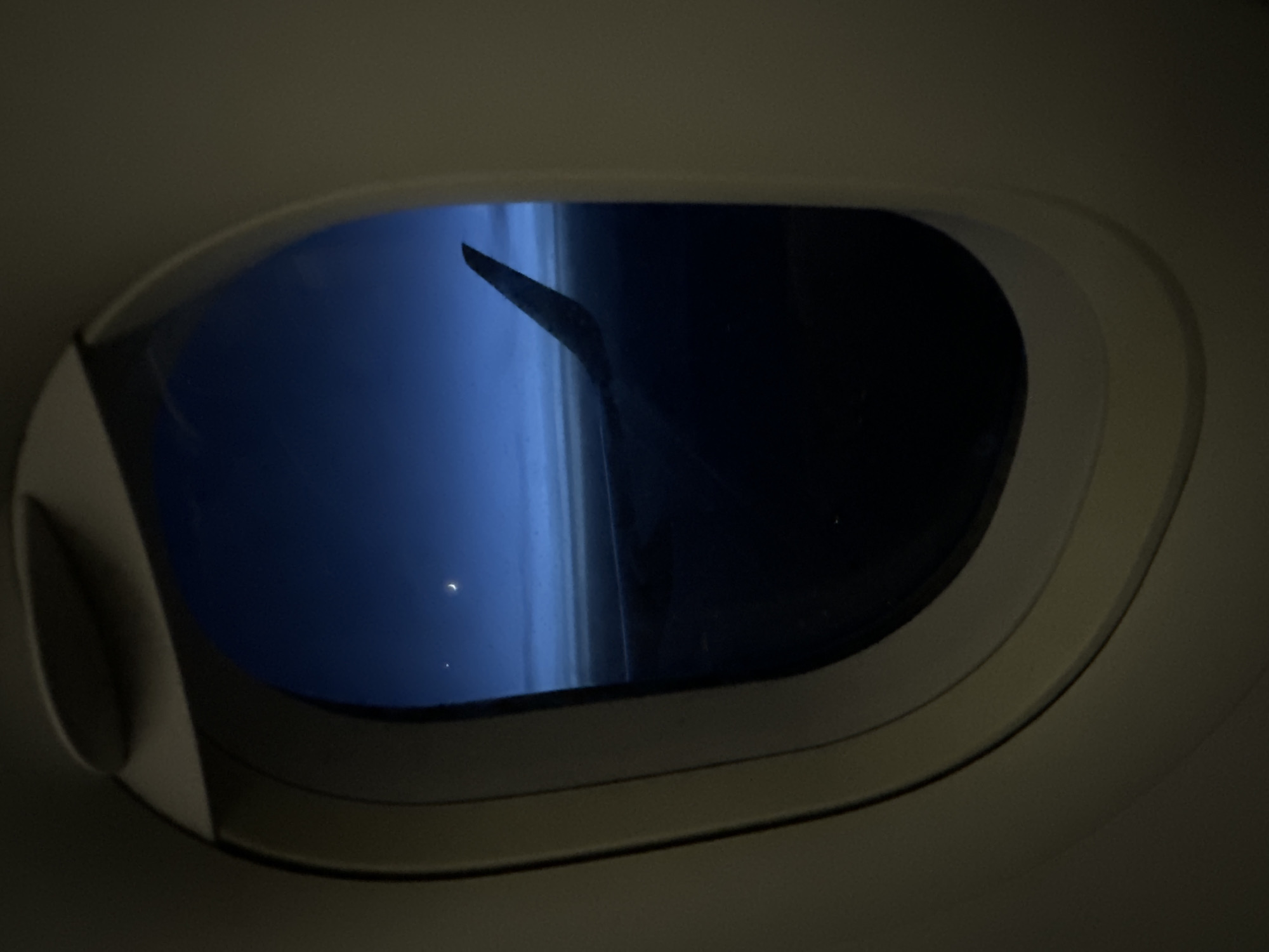 An airplane window provides a view of the night sky. A portion of the wing is visible. The sky is dark blue. Two faint dots of light, possibly stars or other celestial objects, are also visible through the window. The interior of the airplane, specifically the window frame and nearby wall, is muted in color and dimly lit. The location is not discernible from this image.