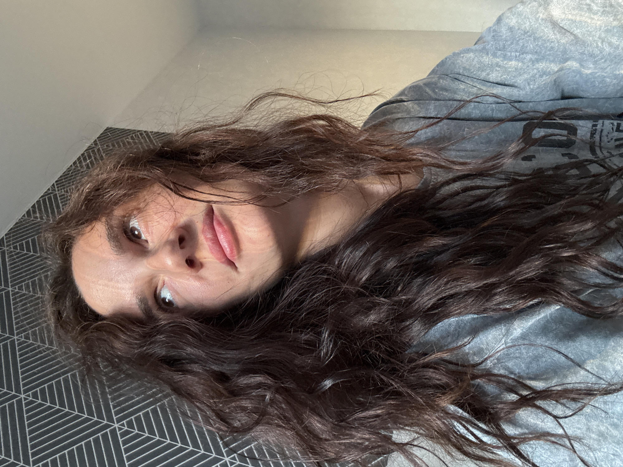 A young woman with long, dark, wavy hair is captured in a medium shot, looking upwards and to the left. She is wearing a gray t-shirt. The background features a wall with a geometric pattern and a plain wall. The scene appears to be indoors, likely a bedroom, in the city of Chaikovsky, Russia.
