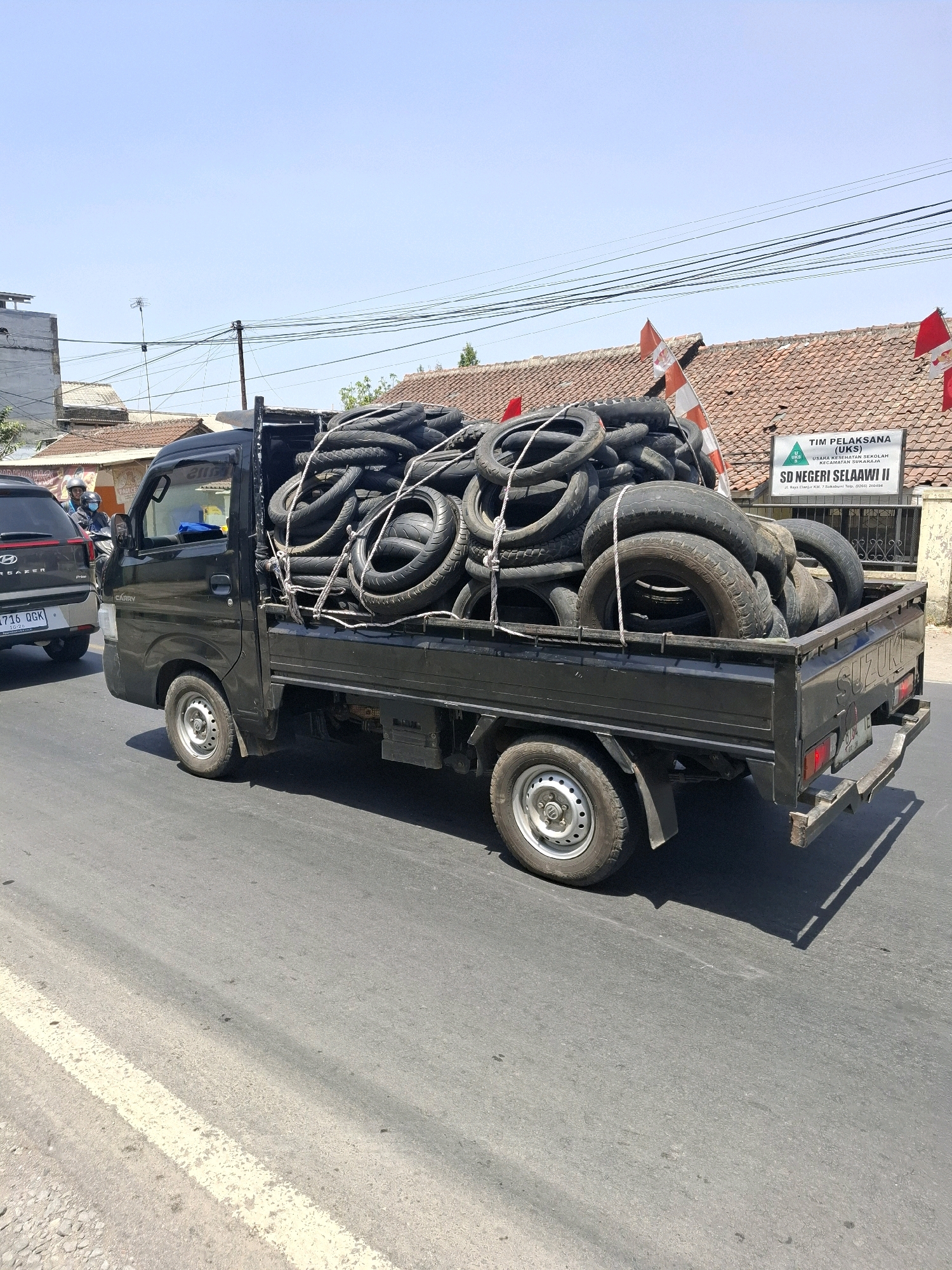 A black Suzuki Carry pickup truck is driving down a paved road. The truck's bed is filled with a large quantity of used tires, which are secured with rope. The truck is in motion, indicated by the slight blur of the tires and road. In the background, there are buildings with tiled roofs, a street sign for "SD NEGERI SELAAWI II," and power lines. A silver car is visible to the left of the truck, and two people on a motorcycle are in front of it. The sky is clear and blue. The image appears to be taken in Selaawi, Indonesia, as suggested by the street sign. The overall scene depicts transportation of recycled materials on a sunny day in a semi-urban environment.