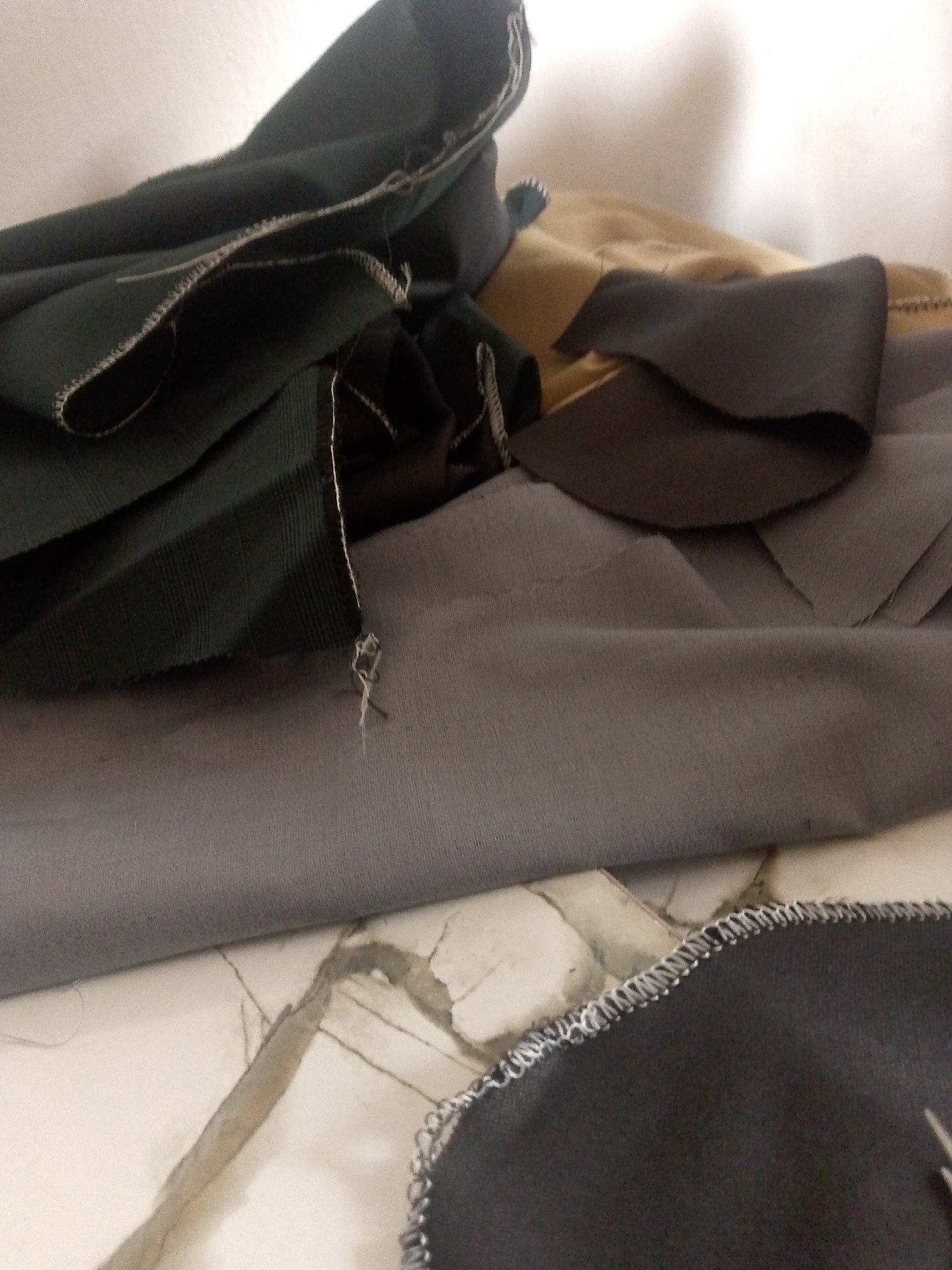 The image shows several pieces of fabric with a visible marbled surface beneath. The fabrics are folded and layered, with some showing overlocked edges. One piece is a dark green fabric with visible vertical ribbing, another appears to be a tan or light brown fabric, and a darker grey fabric is also present. The marbled surface has a pattern of grey and beige streaks. There is no discernible location in the city of Jalingo, Nigeria.