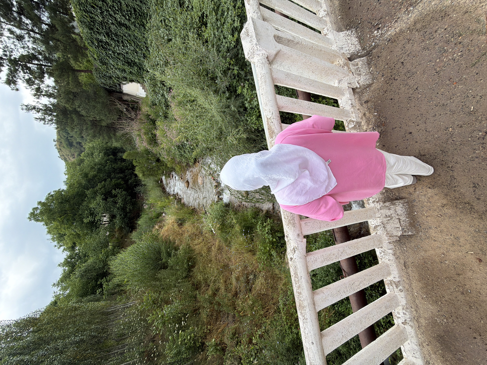 An outdoor, eye-level shot shows a person wearing a pink hijab and a pink tunic, white pants, and white sneakers standing on a bridge. The person is facing away from the camera and looking down at a stream. The stream runs through a rocky, overgrown ravine. The ravine is covered with trees and foliage on both sides. The sky is overcast.
