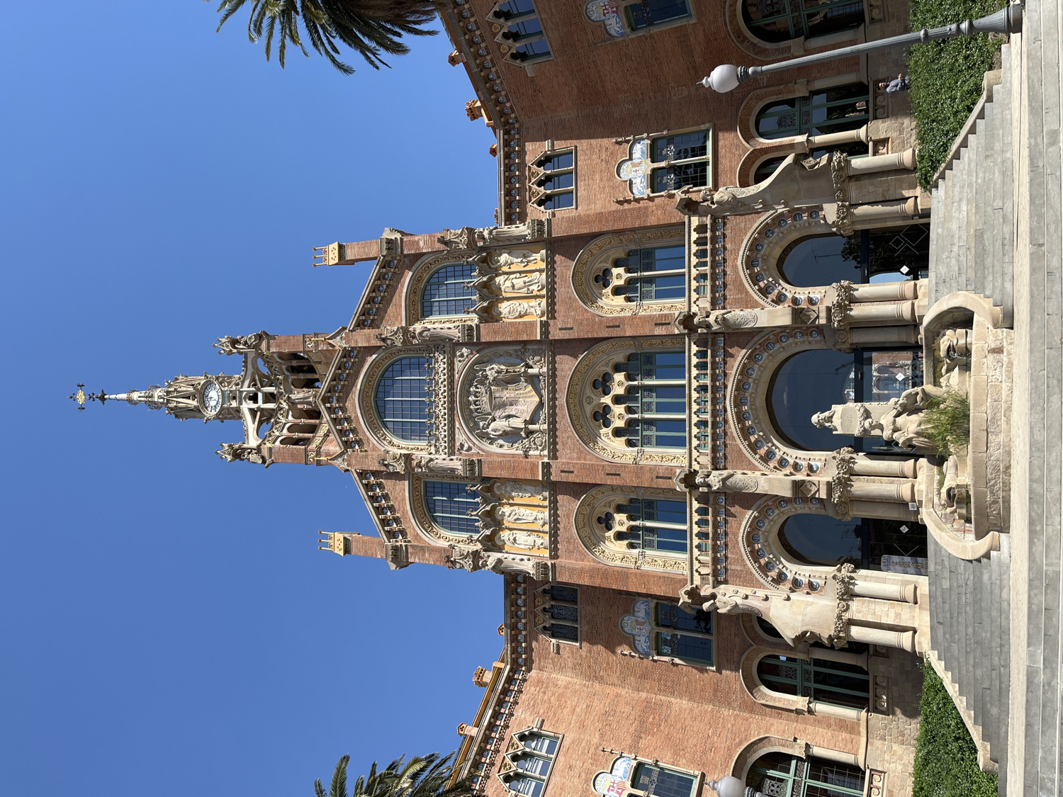 This image captures a close-up of a magnificent, highly ornate building under a clear, bright blue sky, typical of Barcelona, Spain, and strongly resembling the Hospital de Sant Pau. The architectural style is elaborate Modernista, characterized by its intricate details and rich decoration.
The central feature is a tall, slender spire culminating in a decorative finial, with a visible clock face positioned just below its apex. The building's facade is constructed primarily of brick, accented extensively with intricate stone carvings, sculptures, and detailed friezes. Numerous arched windows, some featuring delicate stained-glass patterns and others with decorative grilles, are spread across multiple levels. Statues of figures and other sculptural elements, including gargoyle-like forms, are integrated into the window frames and wall sections, adding to the building's grandiosity.
On the right side of the image, wide stone stairs lead up to an entrance, flanked by lush green bushes and a classic lamppost. The lower section of the building also displays large arched entrances, supported by sturdy columns and adorned with more stone embellishments. A portion of a palm tree is visible in the upper left corner, adding a Mediterranean touch.
The scene is bathed in bright daylight, suggesting it is midday or early afternoon, with strong shadows accentuating the three-dimensional architectural features. No distinct human activity is captured, nor is any legible text visible on the building's facade. The image emphasizes the exquisite craftsmanship and historical significance of the structure.
