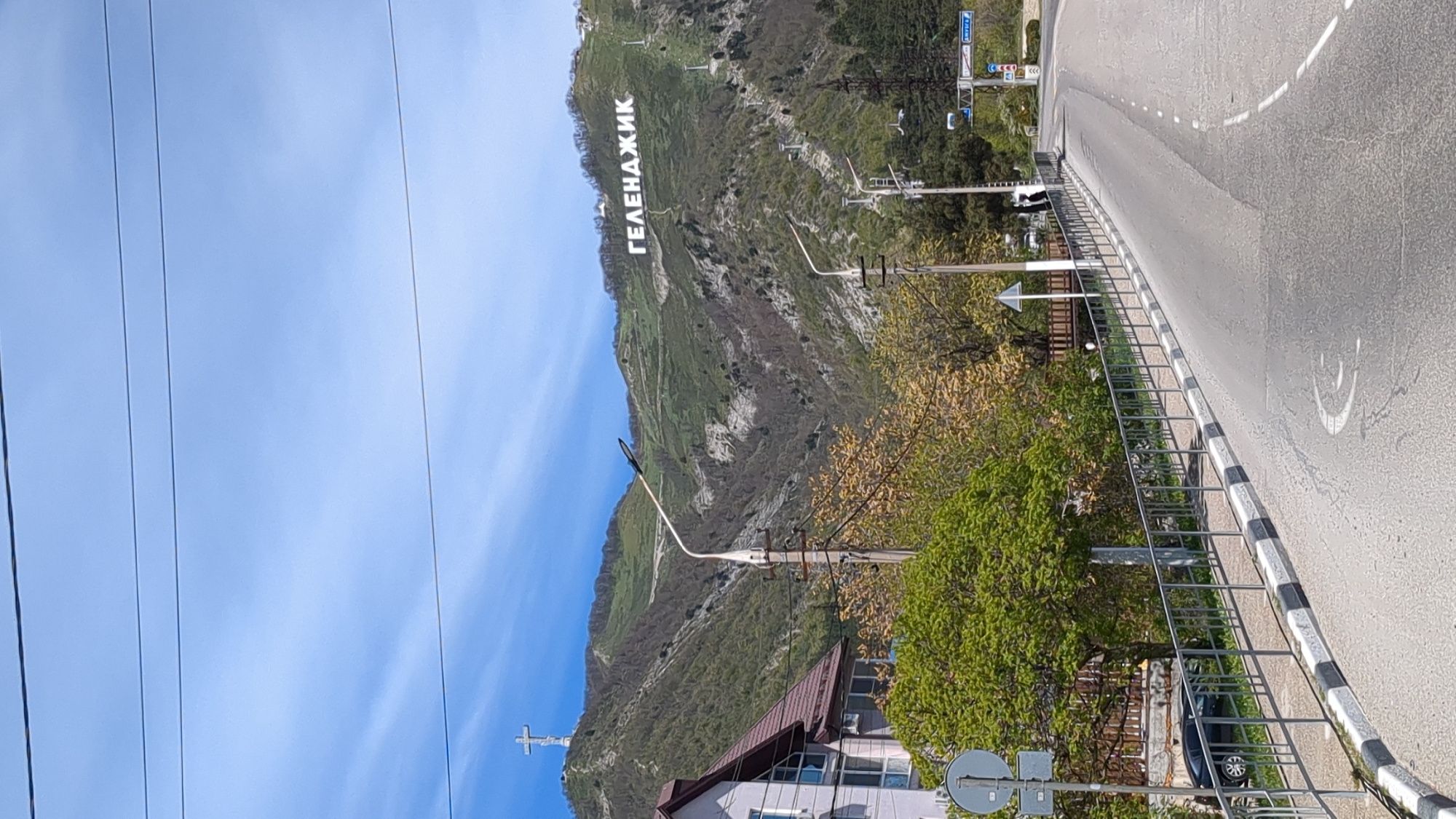 The image depicts a bright, daytime scene in Gelendzhik, Russia, viewed from alongside a road. The most prominent feature is a large mountain in the background, covered in green and brown vegetation, with large white letters spelling "ГЕЛЕНДЖИК" (GELENDZHIK) emblazoned across its slope.
To the right, a paved road curves gently, marked by a dotted white line down its center. A distinct white smiley face is painted on the asphalt. Along the edge of the road, there's a sidewalk separated by a black and white striped curb, and a metal guardrail runs parallel to the road. Further right, several traffic signs are visible, though their text is illegible.
In the mid-ground and foreground, various trees and bushes with developing spring foliage (light green and yellowish leaves) are visible, along with some utility poles and overhead power lines stretching across the sky. Below the main mountain, on a separate, lower peak to the left, a large white cross stands out against the green landscape. Part of a building with a brown roof is visible in the lower foreground, surrounded by trees.
The sky is clear and a vibrant blue, suggesting sunny weather. No specific human activity is depicted, but the scene captures an urban-natural interface along a roadside, likely during late morning or early afternoon given the bright, direct sunlight.