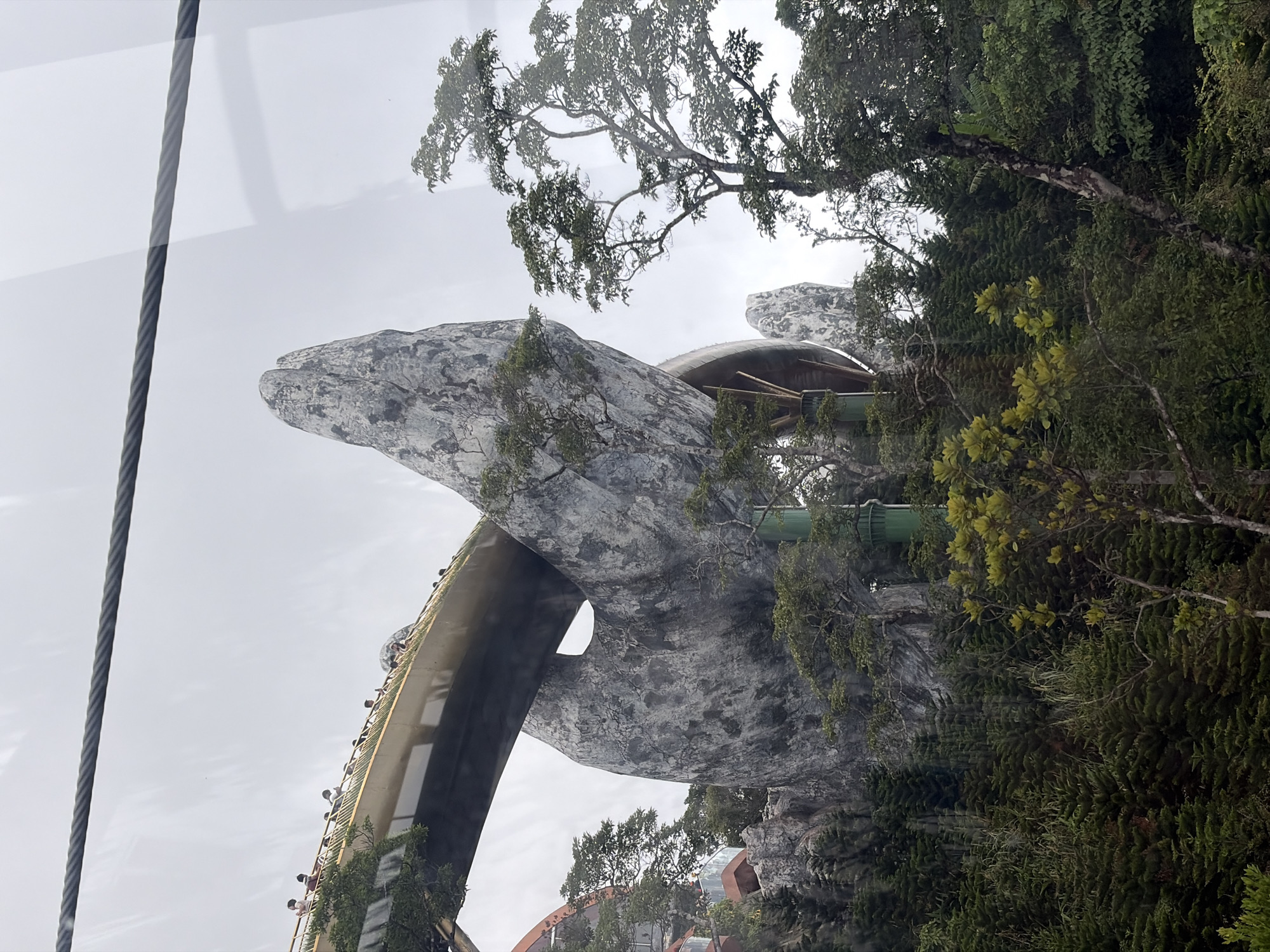 A low-angle shot depicts a structure resembling a massive stone hand or mountain, covered in greenery, viewed from an elevated position with a cable visible at the edge of the frame. The sky is overcast. Located in Bà Nà Hills near Đà Nẵng, Vietnam, this is the Golden Bridge, a pedestrian walkway designed to look like it's held up by giant hands. The walkway is visible along the arm of the structure, dotted with tiny figures of people. The vegetation consists of trees and vines. A metal structure is visible amongst the greenery around the 'hand'. The shot is taken through glass or a window of some kind.
