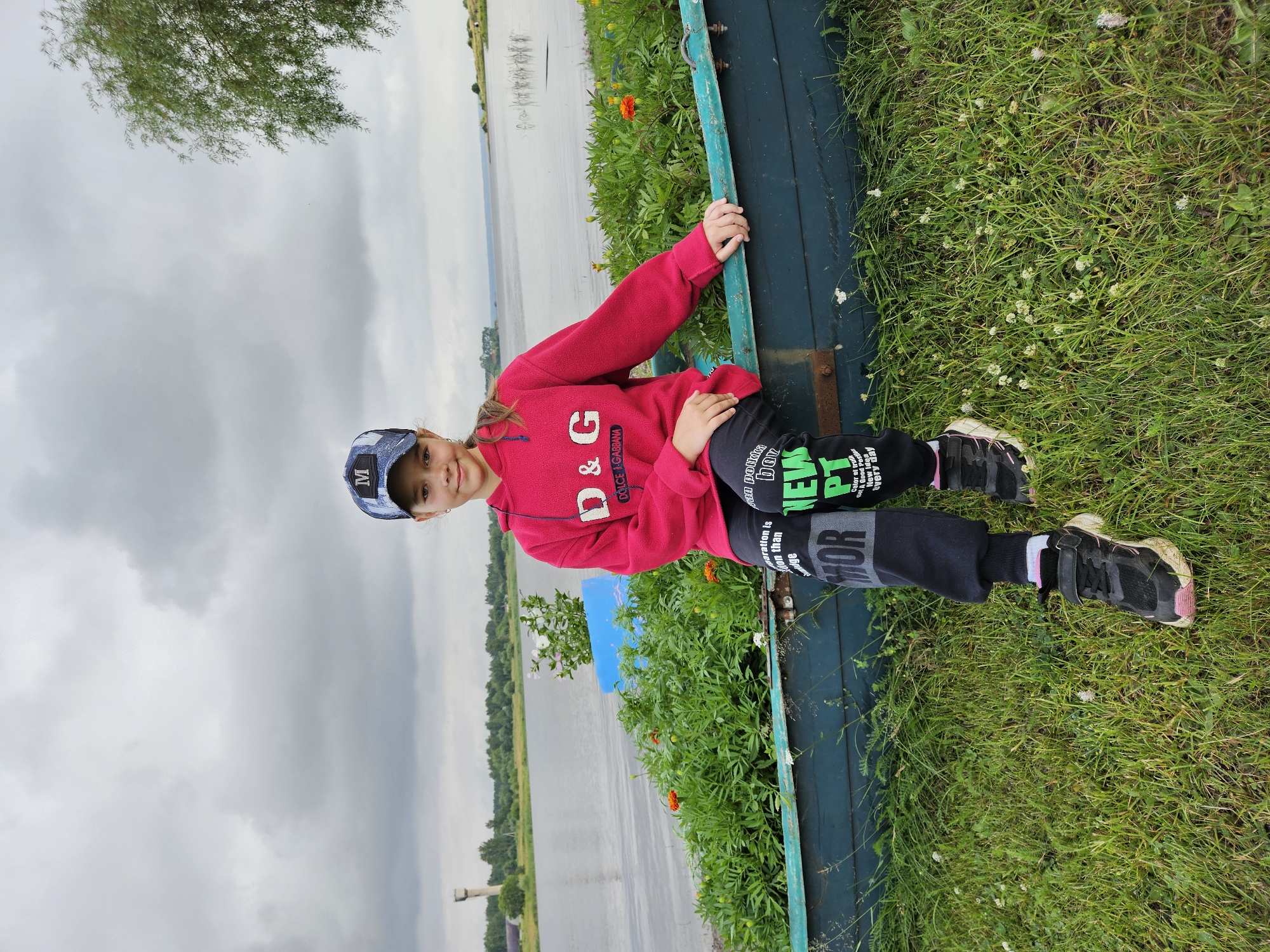 A young girl wearing a pink hoodie and a blue baseball cap with an "M" logo sits on the edge of a blue boat. She is facing the camera and smiling. The boat is positioned on a grassy area next to a body of water, likely a lake or river. Green plants and orange flowers are growing in and around the boat. In the background, a distant tower and trees are visible under a cloudy sky. The overall scene suggests an outdoor, possibly rural, setting. The city of Elovo, Russia, is not depicted in the image, nor is there any indication of it being the location.