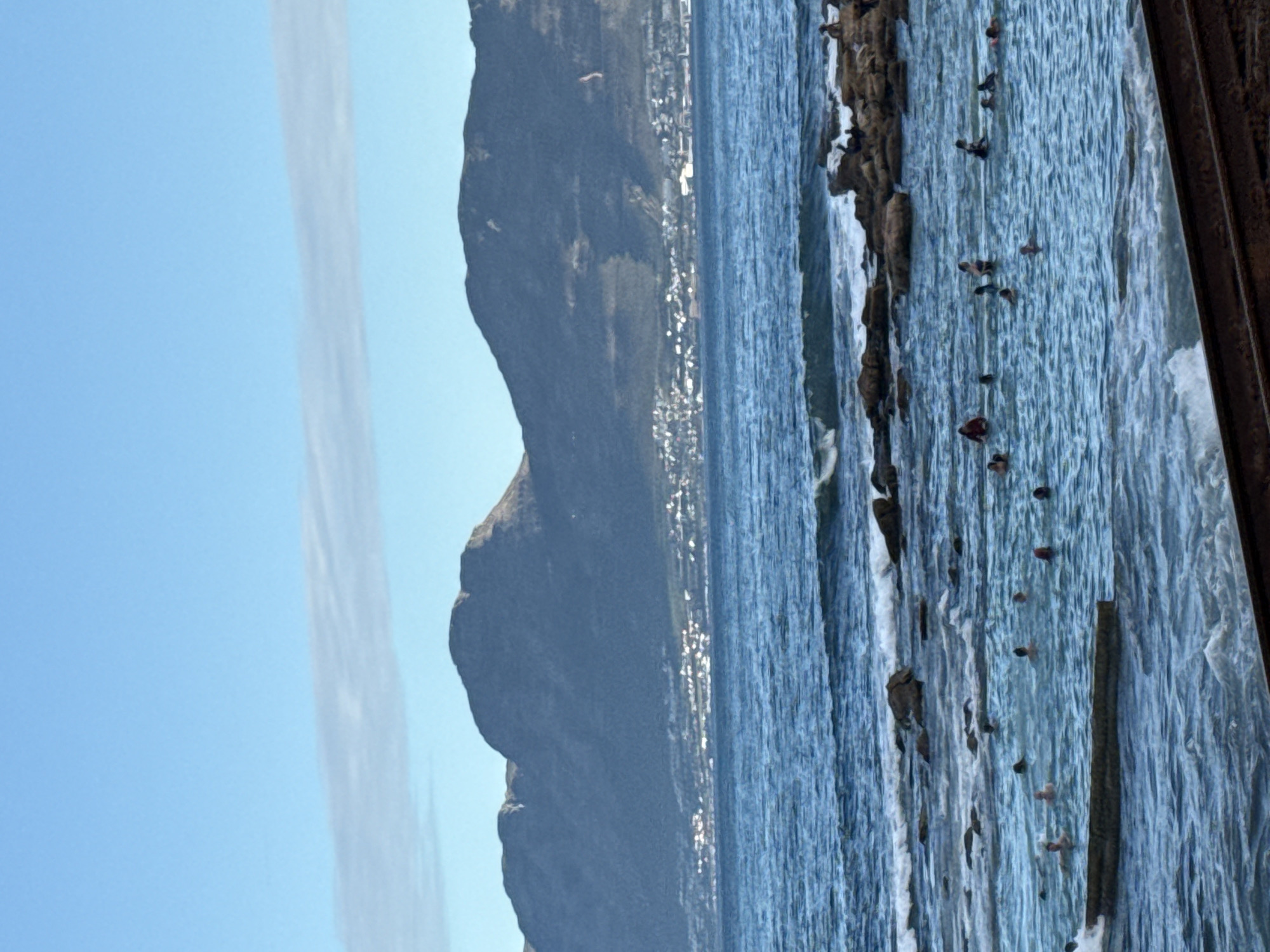 The image shows a coastline scene with a view of a mountain range, likely taken in Cape Town, South Africa, specifically featuring Table Mountain. The sky is clear with some linear cloud formation. The mountain range slopes down towards a city on the coastline. The water appears blue, with waves breaking along a rocky shore. There are people in the water. There is a wooden structure on the right side.
