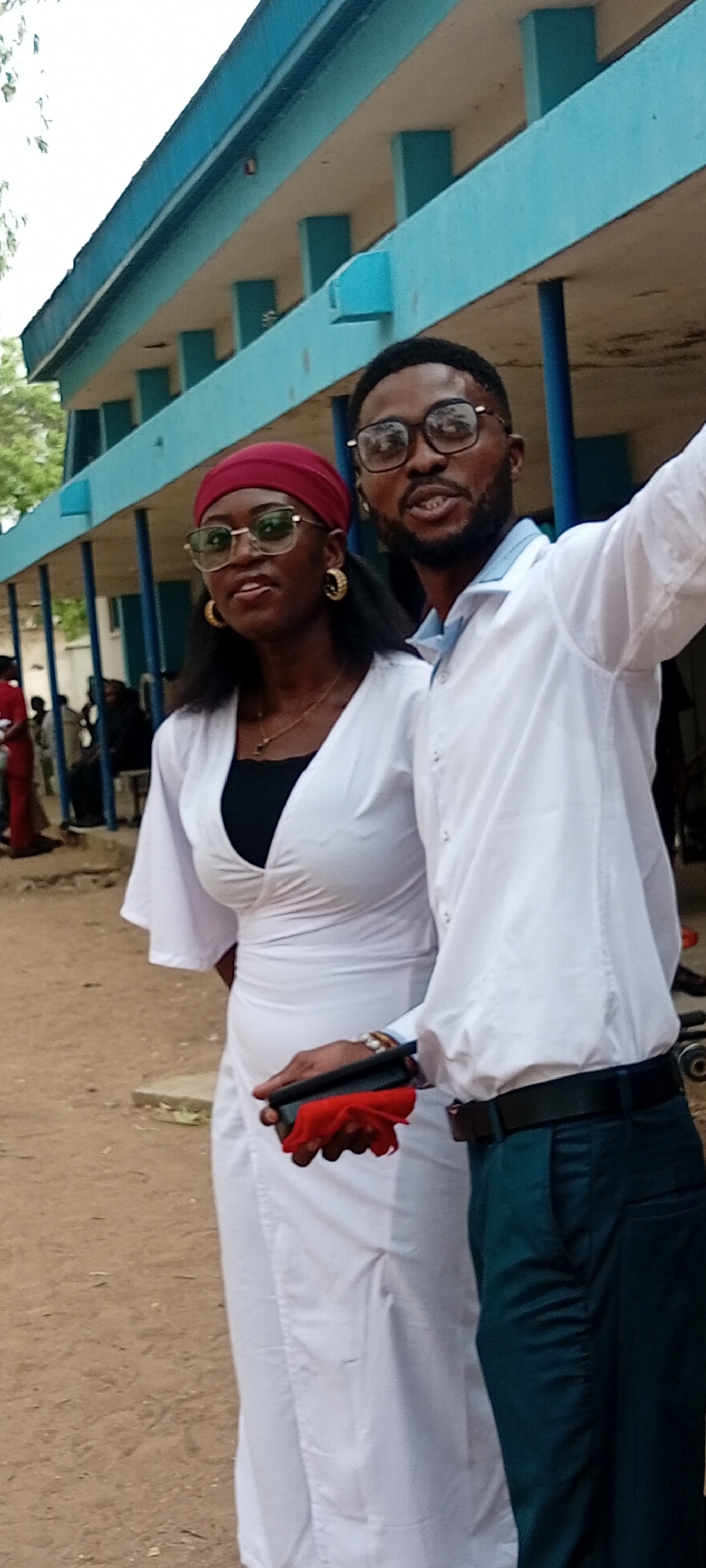 Two young adults pose for a photo in Goron Dutse, Nigeria. The woman wears a white dress, round sunglasses, gold hoop earrings, and a red headscarf. The man wears a white button-down shirt, black-rimmed glasses, dark trousers, and a black belt. The man raises one arm. The woman holds a black object with a red cloth. They are standing in front of a light blue building with pillars. Other people are visible in the background. The ground is dirt or sand.