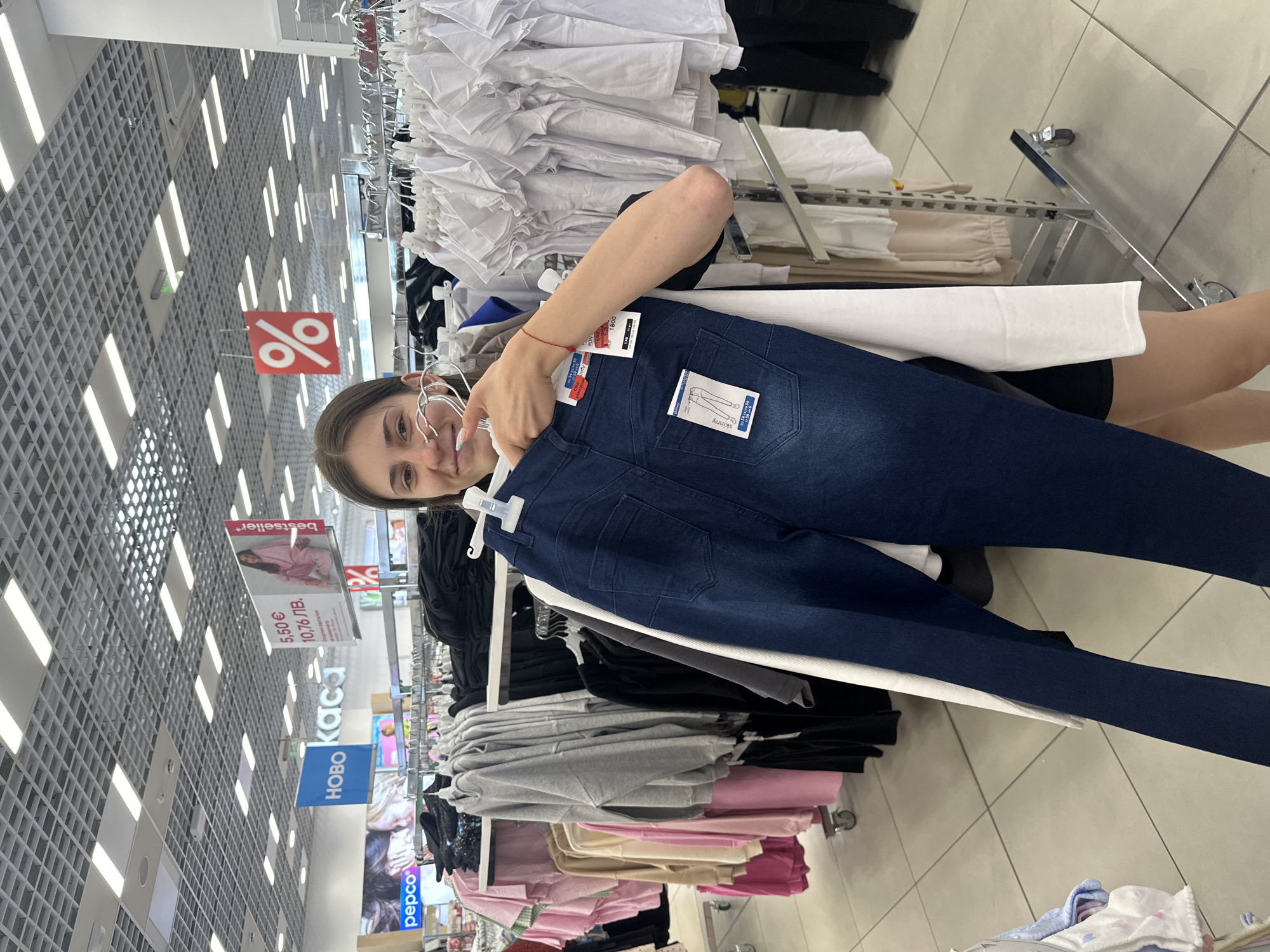 A young woman is holding a pair of dark blue jeans in a clothing store. She is standing in front of racks of clothes, including white t-shirts and pink and beige tops. There are signs in the background with Bulgarian text and a percentage sign indicating a sale. The store appears to be a large retail establishment with a tiled floor and a suspended ceiling with lighting. The woman is smiling and looking at the camera. The image was taken in Panagyurishte, Bulgaria.