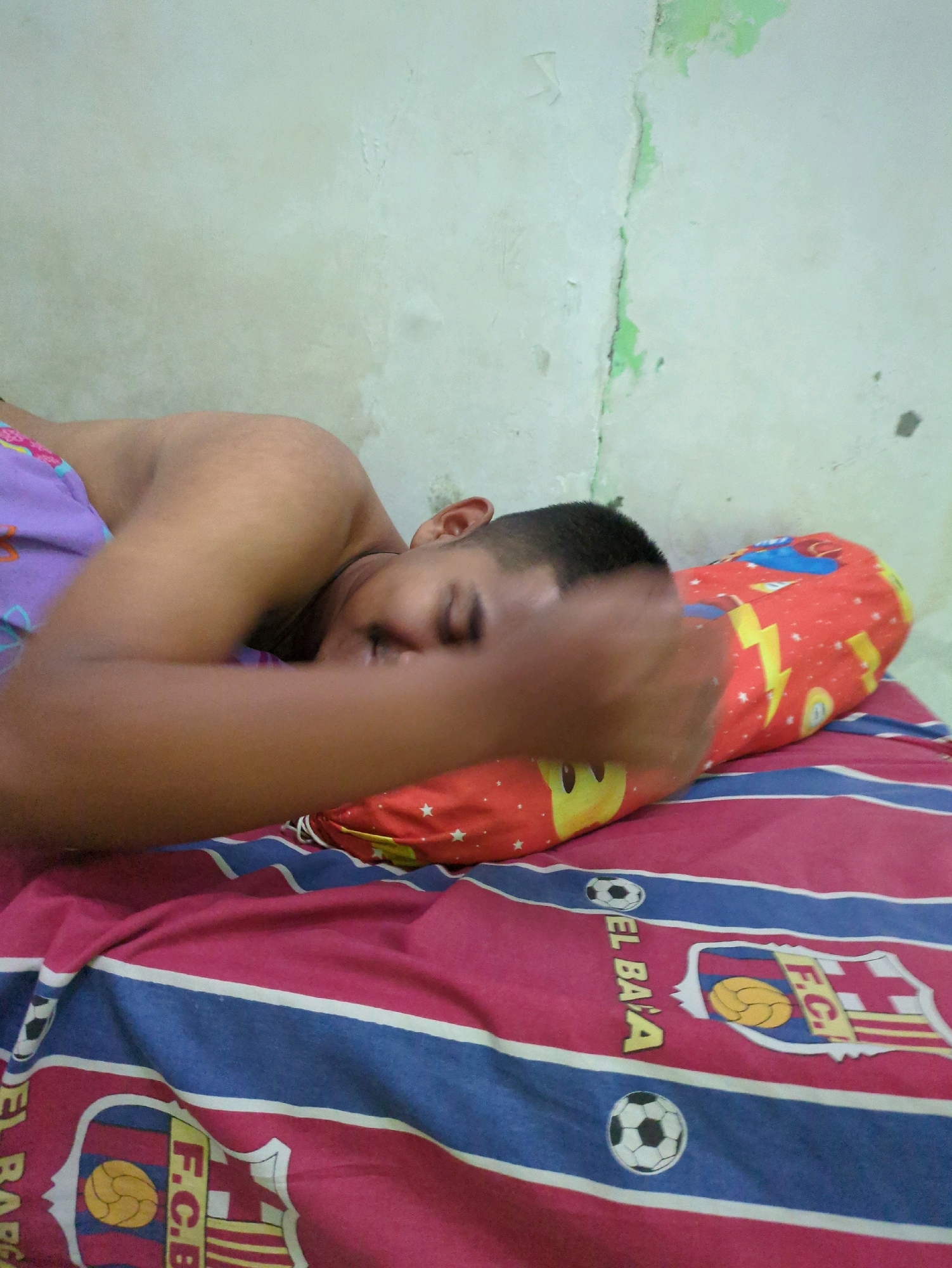 A young male is sleeping on a bed in a bedroom. The bed has a red and blue striped blanket with football club logos. A red pillow with yellow lightning bolts and cartoon characters is under his head. The wall behind the bed is off-white with green paint peeling off in places. The scene appears to be in a residential setting.
