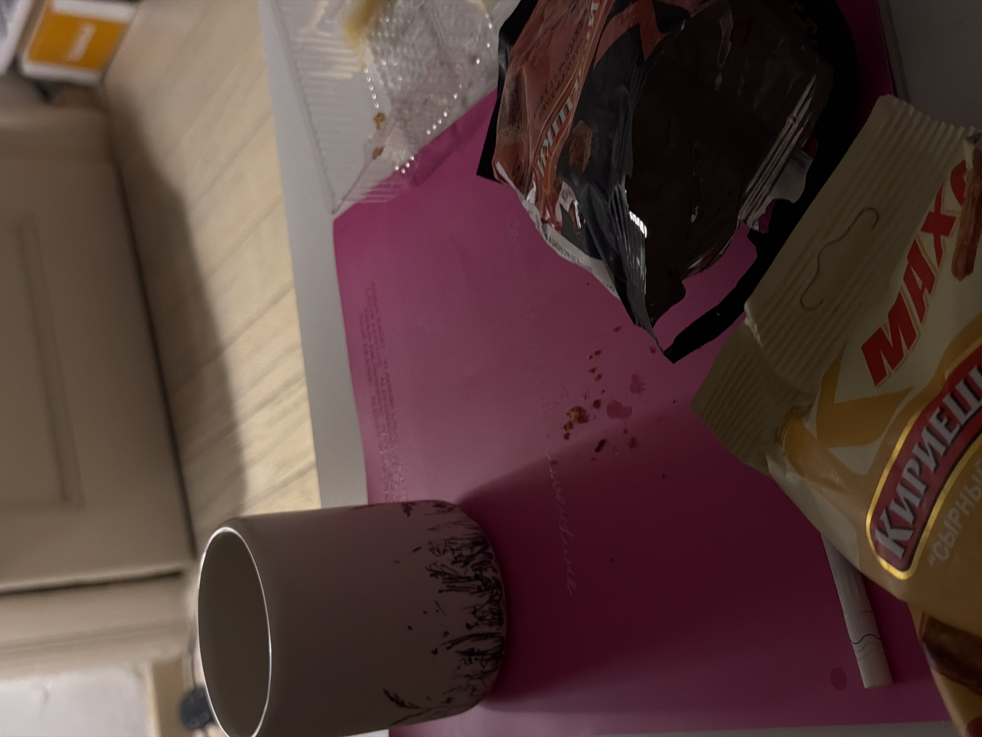 A close-up, overhead shot depicts a casual, indoor scene, likely a desk or table surface. A pink notebook lies open, its surface slightly messy with crumbs and what appear to be dried liquid droplets. A white mug with a dark, grass-like pattern near its base sits to the left, casting a shadow on the notebook. In the foreground, a package of "Kirieshki" (Russian snack) and another partially opened snack bag are visible. A single cigarette lies next to the "Kirieshki" package. The background is out of focus, showing a wooden floor and what appears to be a door or wall. The lighting is dim, creating shadows and highlighting textures. The scene suggests a moment of rest or a casual break, possibly in a domestic setting or a study area. There is no indication of the city of Vracar Urban Municipality, Serbia, or any specific location details beyond what is visually present in the image.