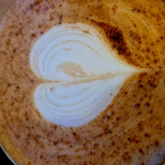 The image is a close-up, top-down shot of latte art in a cup of coffee. The central feature is a distinct heart shape, expertly formed in white milk foam, set against a rich, warm brown and orange background of coffee and frothed milk.
The heart itself is smooth and creamy white, exhibiting subtle swirling patterns within its form, suggesting the delicate technique of latte art. The edges of the heart are clearly defined, contrasting sharply with the darker liquid around it.
The background, which comprises the rest of the coffee surface, is a blend of deep golden-browns and burnt oranges. It has a slightly textured, frothy appearance. Scattered across this darker surface are numerous tiny, reddish-brown specks, likely cinnamon, cocoa powder, or fine coffee grounds, adding to the visual texture and depth of the drink. Some areas of the background also show faint swirling patterns, indicating the movement of the milk and coffee.
The overall impression is one of a freshly prepared, inviting, and aesthetically pleasing beverage. The lighting appears soft and even, highlighting the contrast between the white foam and the darker coffee. There are no other objects, people, text, or discernible environment visible in the image, keeping the focus entirely on the latte art.