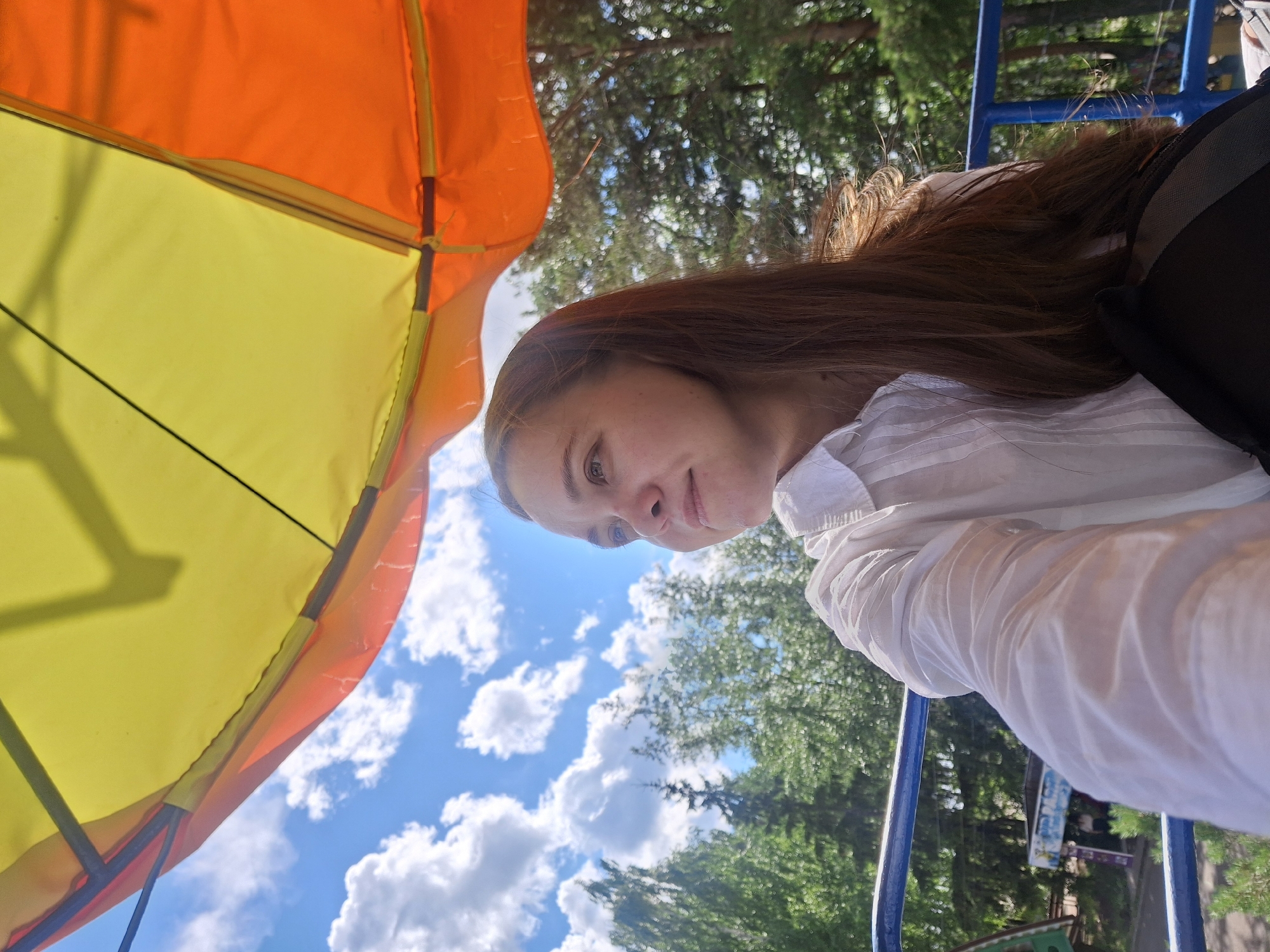 A woman with long, reddish-brown hair is captured in a selfie, her face turned towards the camera. She is wearing a white, collared shirt with subtle pleating on the sleeves and front. Behind her, a large, bright yellow and orange umbrella or canopy dominates the left side of the frame, casting shadows. The sky above is a clear blue, dotted with fluffy white clouds. Green trees and foliage are visible in the background, suggesting an outdoor setting on a sunny day. A blue metal railing, possibly part of playground equipment or a structure, runs along the right side of the image. The woman's expression is relaxed and pleasant, with a slight smile. The image appears to have been taken outdoors during daylight hours. The location context provided is Chaikovsky, Russia. There is no visible text in the image.