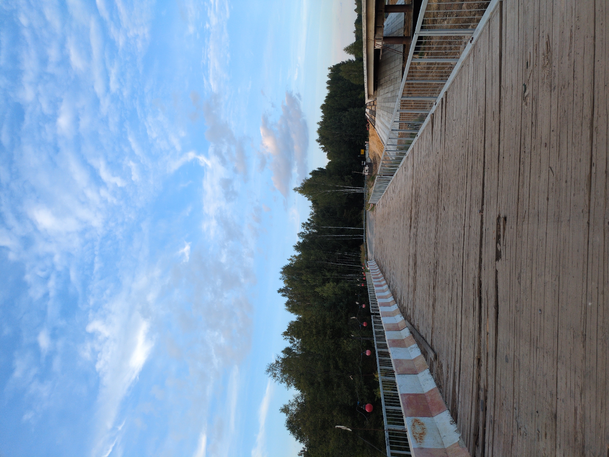 A low-angle, full shot captures the perspective of someone standing on a wooden bridge, looking forward. The bridge is constructed of weathered wooden planks laid horizontally, with a metal railing along the right side. The railing is topped with a concrete barrier painted with alternating red and white bands. Beyond the bridge on the right, an unfinished concrete bridge structure is visible, supported by large cylindrical pillars and surrounded by construction materials and bare earth. On the left side of the wooden bridge, a line of red spherical lights hang suspended by wires above the railing. The bridge curves slightly to the right as it recedes into the distance. A dense, dark green forest with varying tree heights forms a backdrop on the left and in the distance, with a few lighter-barked trees interspersed. The sky above is a bright blue, partially filled with scattered, wispy white clouds. The overall scene suggests a rural or semi-rural setting, possibly indicating ongoing infrastructure work.