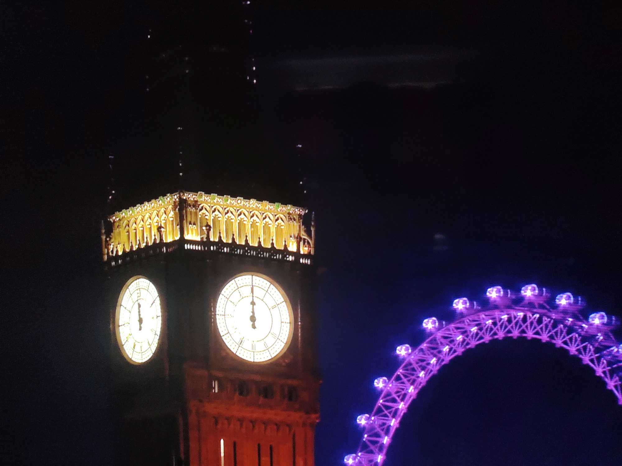The image depicts a night scene in London, United Kingdom, featuring the iconic Elizabeth Tower (commonly known as Big Ben) and a segment of the London Eye. The Elizabeth Tower, illuminated with golden lights, shows two clock faces; the left clock face displays the time as approximately 12:07, and the right clock face is also visible, indicating a similar time. The structure of the tower is intricate with decorative elements at its top. To the right of the tower, a portion of the London Eye is prominently visible, illuminated with vibrant purple lights. The London Eye appears as a large Ferris wheel with numerous cabins. The background is dark, suggesting nighttime, with subtle hints of clouds in the upper portion of the frame. Small, bright spots are scattered in the sky above the Elizabeth Tower, which could be distant lights or perhaps fireworks. The image is slightly blurry, particularly in the sky and the details of the London Eye. There are no people or identifiable actions depicted in the scene.