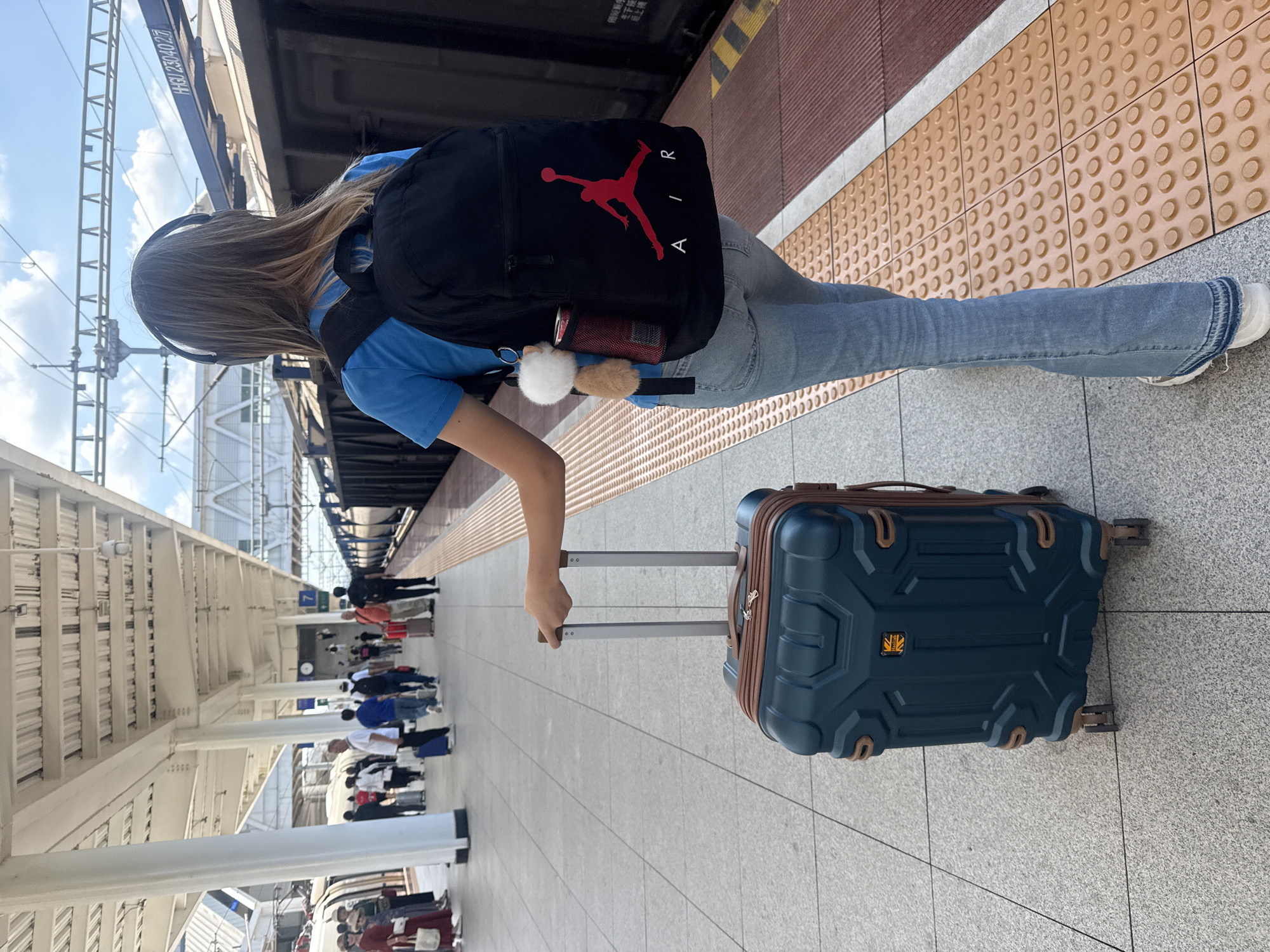 A young woman with blonde hair, wearing a blue t-shirt, blue flared jeans, and white sneakers, stands on a train platform in Wuxing District, China. She has headphones on and is carrying a black backpack with a red Jordan logo. In her right hand, she holds the handle of a dark teal suitcase with brown accents. The suitcase has four wheels and a textured design. Other passengers are visible in the background on the platform, some with their own luggage. A train is parked alongside the platform. The platform is paved with grey tiles and bordered by a yellow textured strip. The scene is under a covered structure with white pillars and a metal roof. The sky is visible above the train and the platform structure.