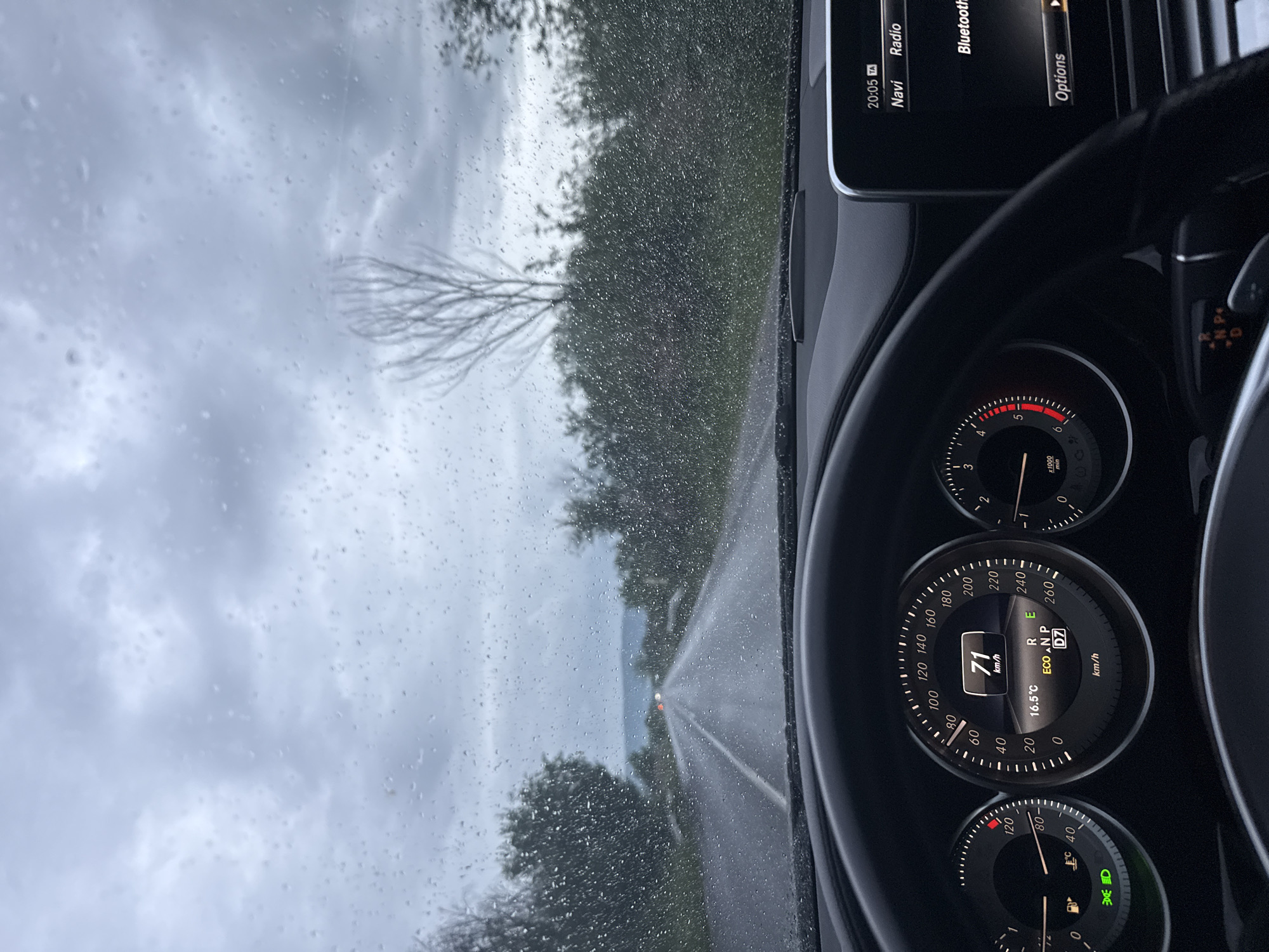 The image is taken from inside a car, likely the driver's seat, showing a rainy day view through the windshield. The focus is on the dashboard and the road ahead. The dashboard displays a speedometer reading of 71 km/h and a temperature of 16.5°C. The car is in "Eco" mode. Outside, the road is wet and surrounded by trees on both sides. The sky is overcast and gray. Based on the weather and roadside vegetation, it could be inferred to be somewhere rural, but without specific landmarks, pinpointing the exact location within Dolna Mahala, Bulgaria, is not possible. The road appears straight and narrow, with a distant light visible further down.