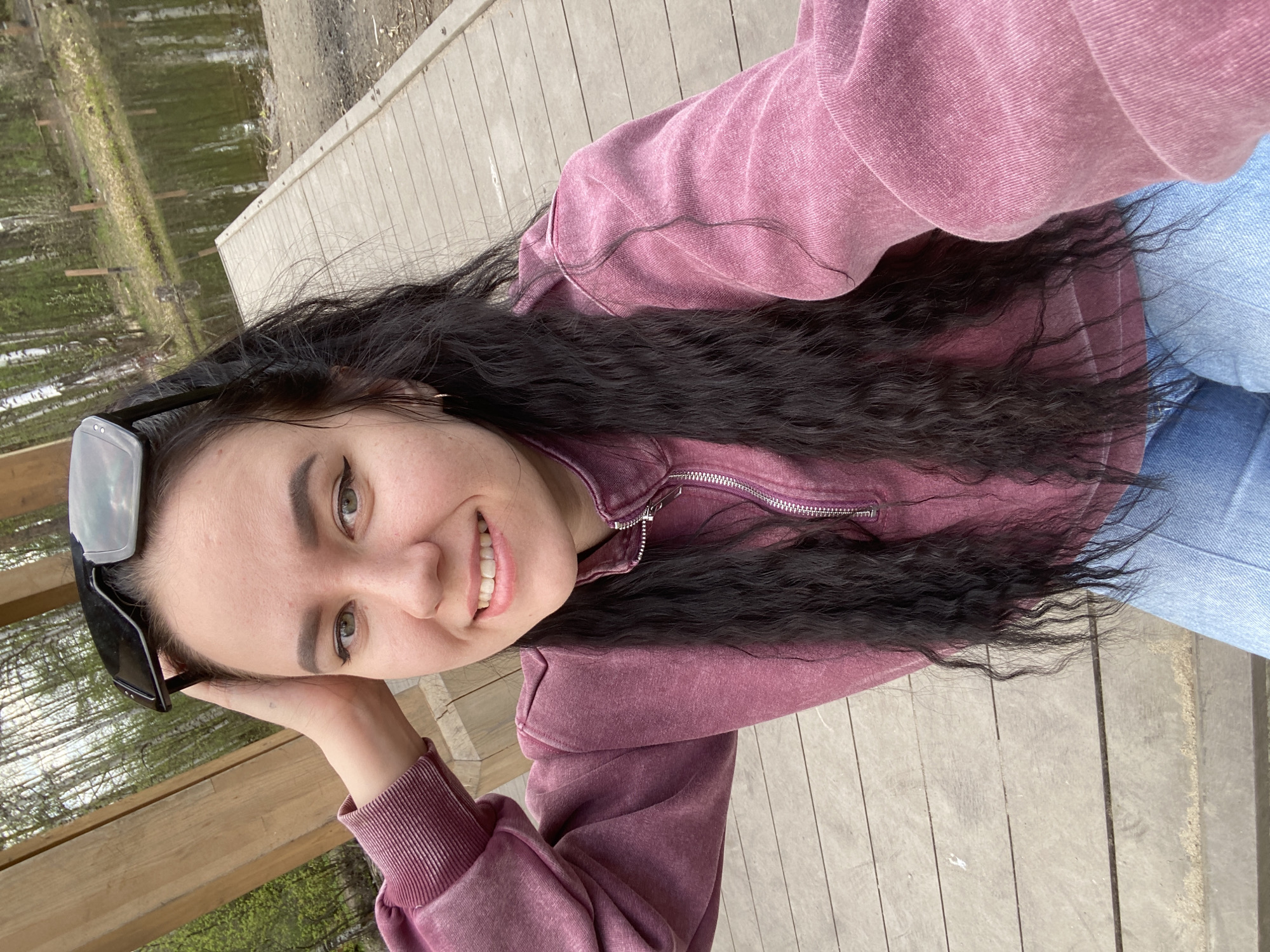 A selfie of a young woman, likely taken outdoors, is prominently featured. She is wearing a mauve-colored zip-up sweatshirt and blue jeans. She has dark, wavy hair and is wearing sunglasses perched on her head. The background suggests an outdoor setting with wooden planks, possibly a boardwalk or deck, and greenery visible in the distance, possibly indicating a park or wooded area. The presence of trees and water suggests a natural or recreational setting. Although the city of Perm, Russia is mentioned, there is no definitive evidence in the image itself to confirm the location.