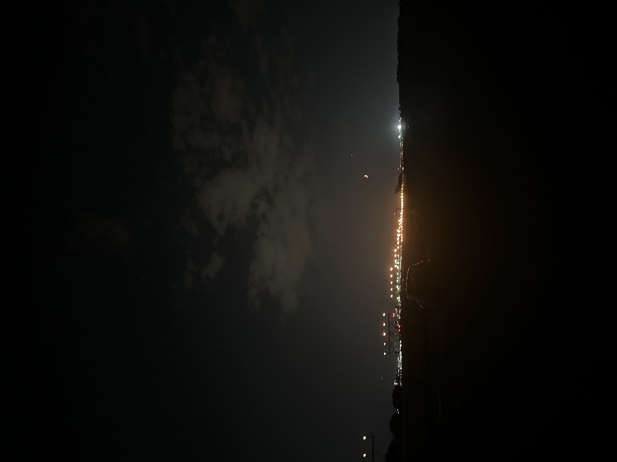 The image depicts a nighttime scene of an airport runway and its surrounding area. In the foreground, a dark silhouette of a car is visible on the right side. The middle ground is dominated by the illuminated runway lights, appearing as a string of bright orange and white orbs stretching across the horizon. Beyond the runway, distant city lights create a glow against the dark sky, suggesting the presence of an urban area. In the upper portion of the sky, scattered clouds are present, with a faint crescent moon and a small airplane visible. The overall impression is of an active airport at night, with the lights of the runway and the city creating a contrasting spectacle against the dark sky. The location is not specified in the provided image, and therefore it cannot be identified as being in Sofia, Bulgaria.