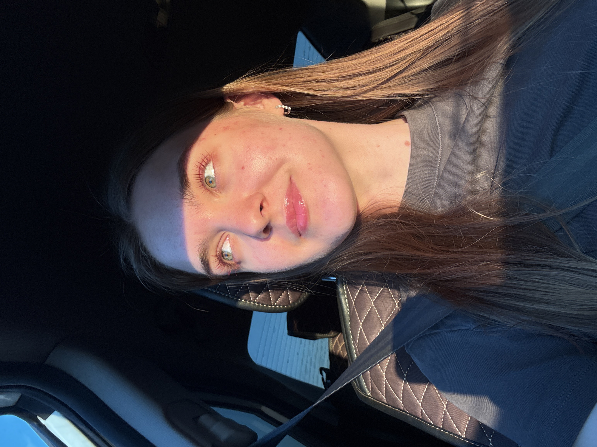The image shows a young woman sitting in the driver's seat of a car, looking out the window. The car's interior is visible, including a seatbelt, quilted seat upholstery, and the dashboard. The woman has light-colored eyes, pink-tinted eyelashes, and is wearing a gray t-shirt and a small hoop earring. Sunlight is casting a warm glow on her face, highlighting her features and creating a shadow across her forehead and right cheek. The background outside the car window appears to be a landscape with a blue sky. The image does not provide any specific information about the location in the city of Ustinovo, Russia.