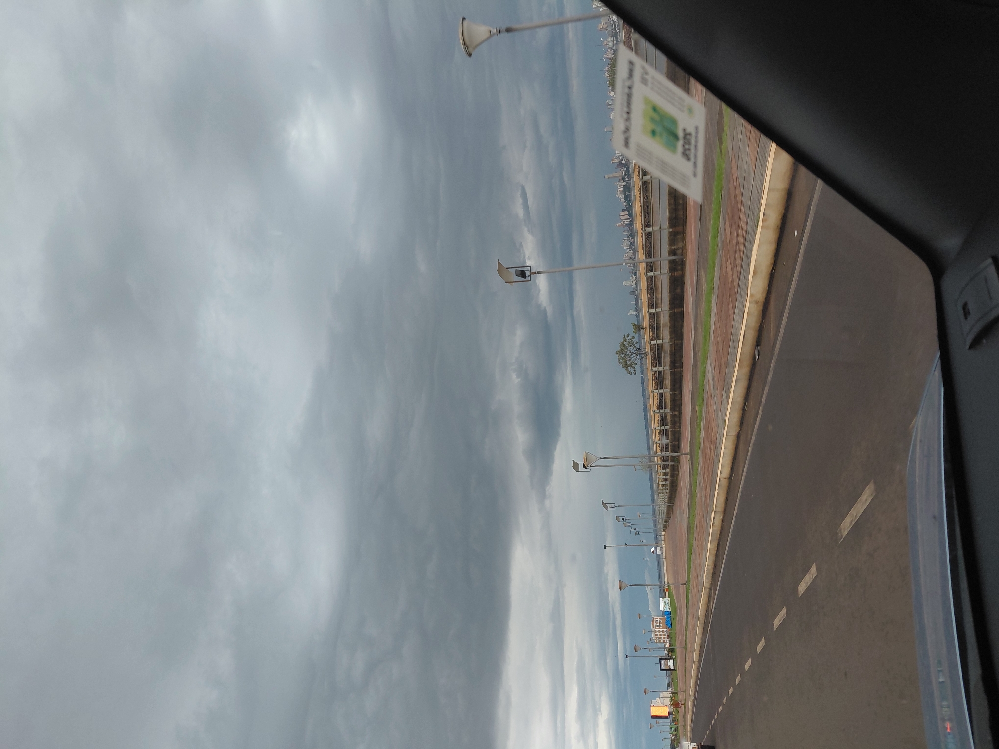 The image captures a view from a vehicle traveling along a road adjacent to a waterfront promenade in Encarnación, Paraguay. The sky is overcast with heavy, dark clouds, suggesting impending rain or a stormy atmosphere. Along the promenade, there is a paved walkway with a series of modern streetlights, a low railing, and planters with grass. In the distance, a cityscape with several tall buildings is visible across a body of water. A sticker with green and white graphics is affixed to the vehicle's windshield in the lower right corner of the frame. The road itself has dashed white lane markings. The scene appears to be in the early stages of twilight.