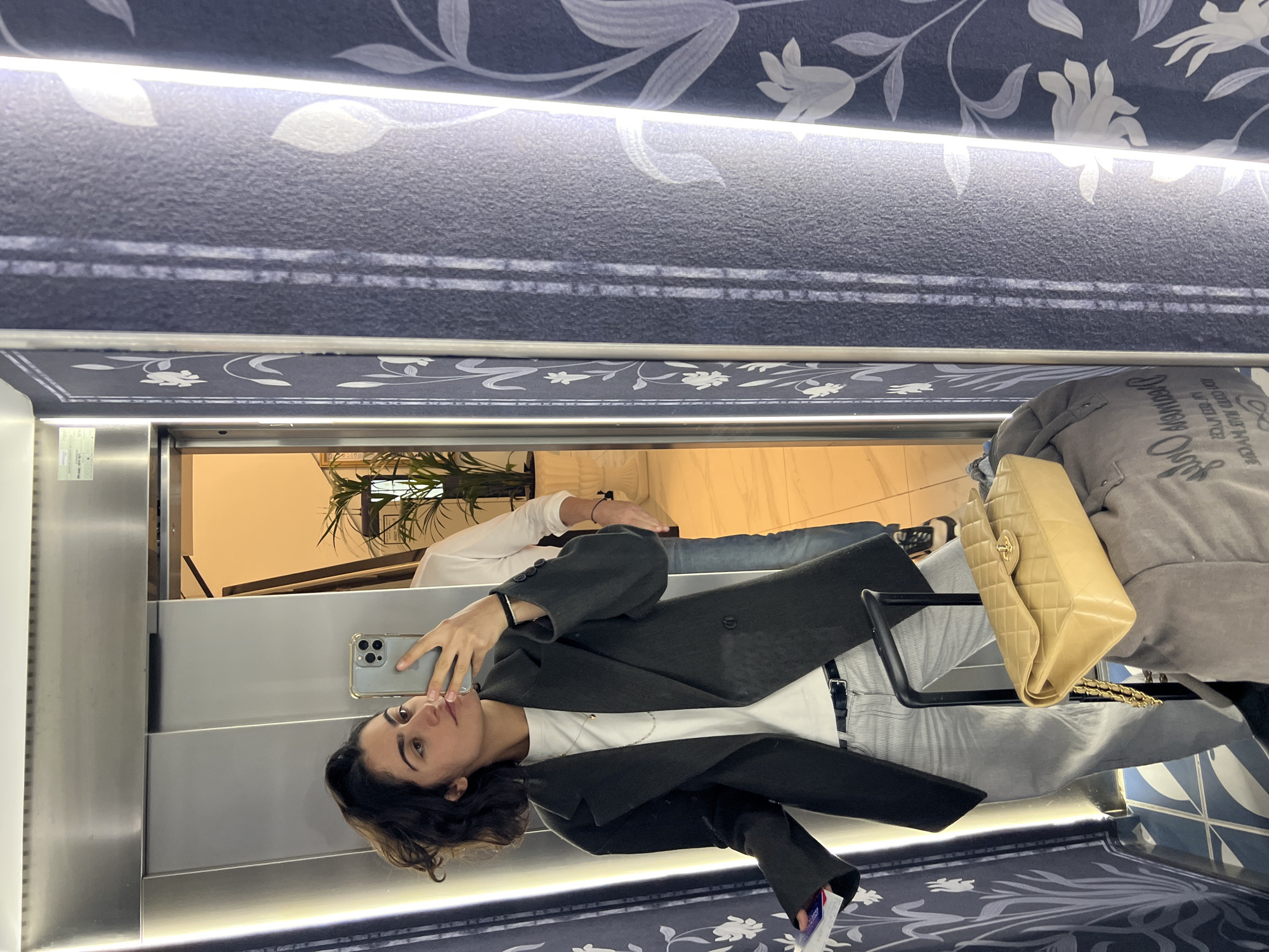 A woman is taking a selfie in an elevator with a patterned wall in Santa Eulària des Riu, Spain. She is wearing a dark blazer over a white top, light-colored pants, and is holding a smartphone. A purse and another person’s arm are visible in the reflection. The elevator interior includes plants and a light-colored floor.