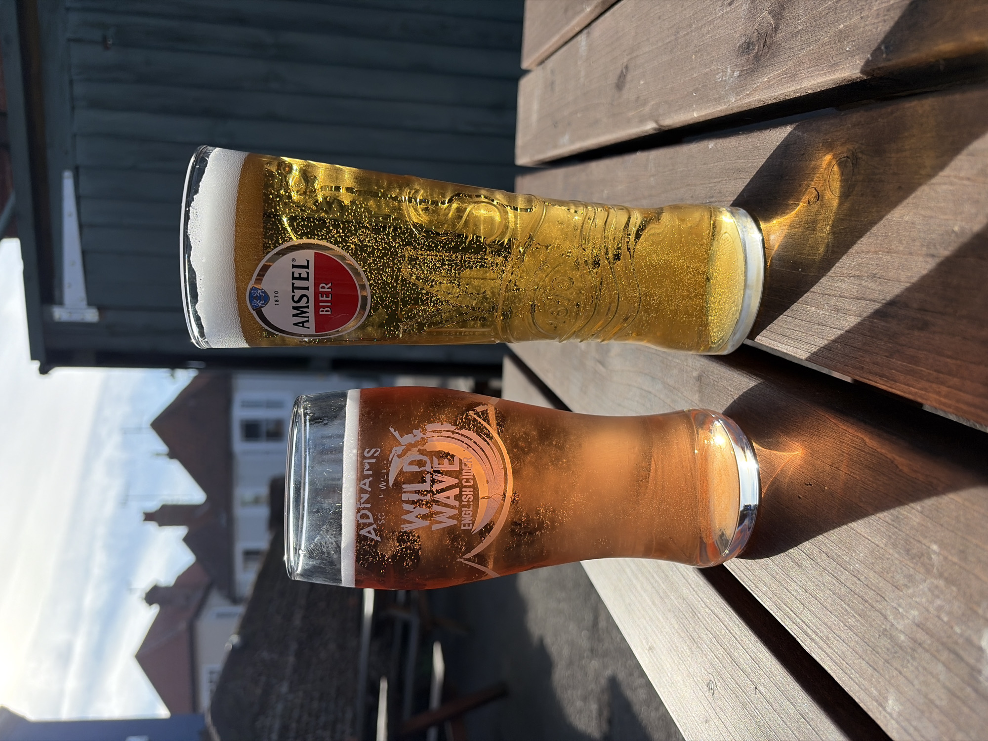 Two glasses, one containing Amstel beer and the other Adnams Wild Wave English Cider, rest on a wooden surface. The Amstel beer glass is positioned at the top of the frame, while the cider glass is below it. Both cast shadows on the wooden surface. In the background, rooftops of buildings are visible, indicating an urban or suburban setting. The location is likely in East Suffolk, United Kingdom. The scene is bathed in sunlight.